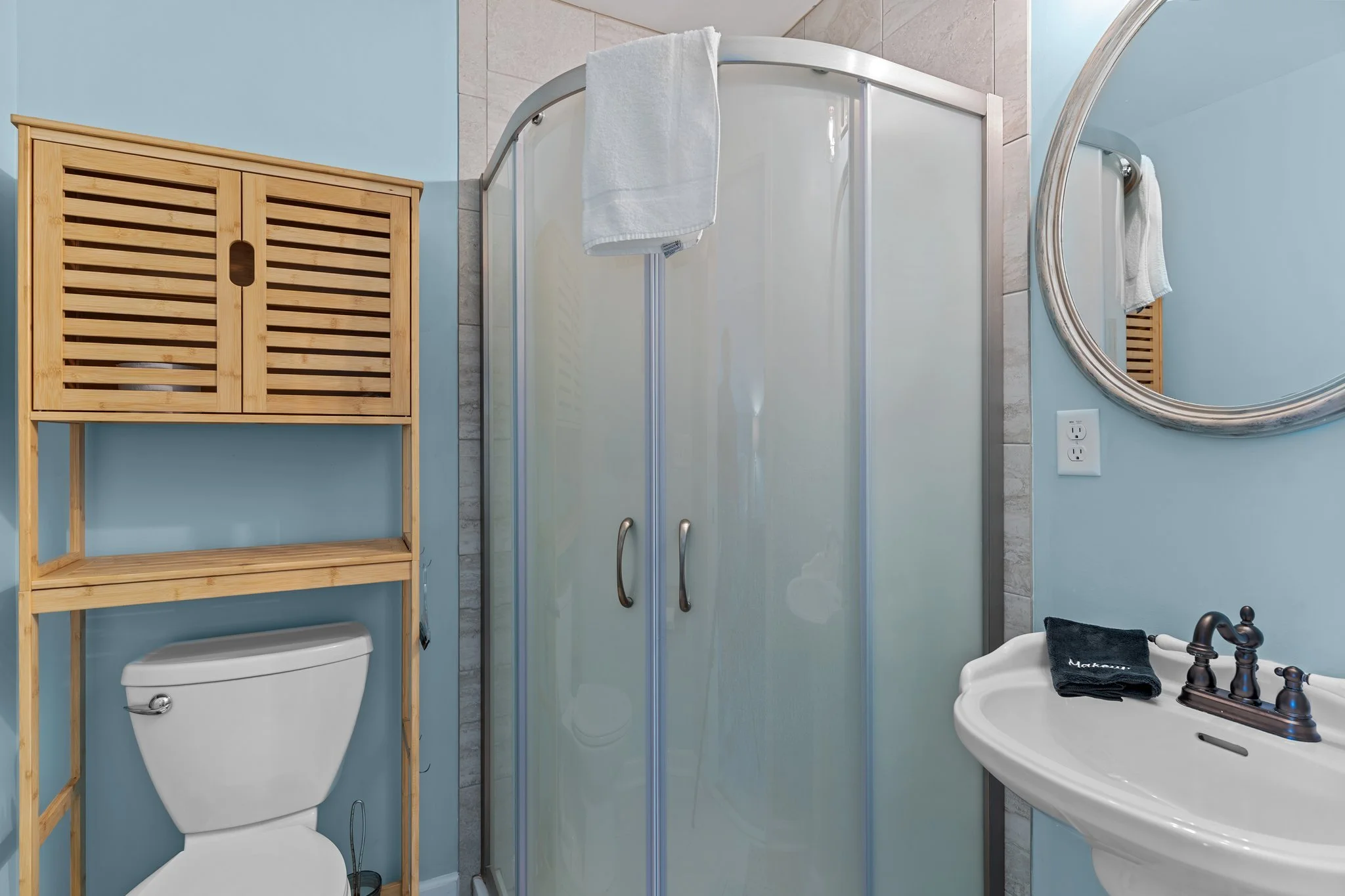 Bathroom with light blue walls, a corner shower with frosted glass doors, a wooden storage shelf above a toilet, and a white sink with black fixtures and a black towel.