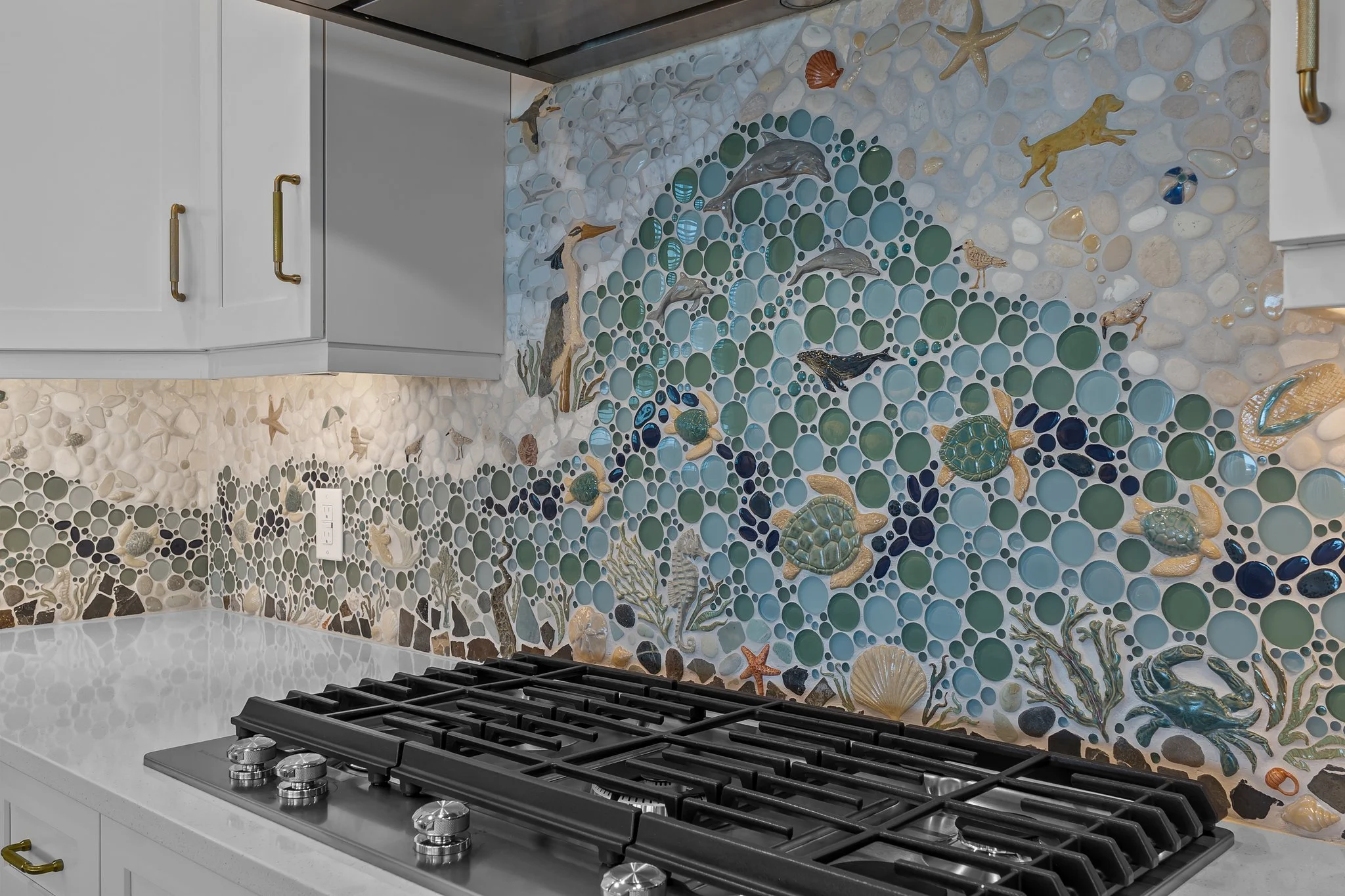 Kitchen backsplash with sea life artwork, featuring dolphins, fish, turtles, and shells, surrounding a gas stove with white cabinetry and gold handles.