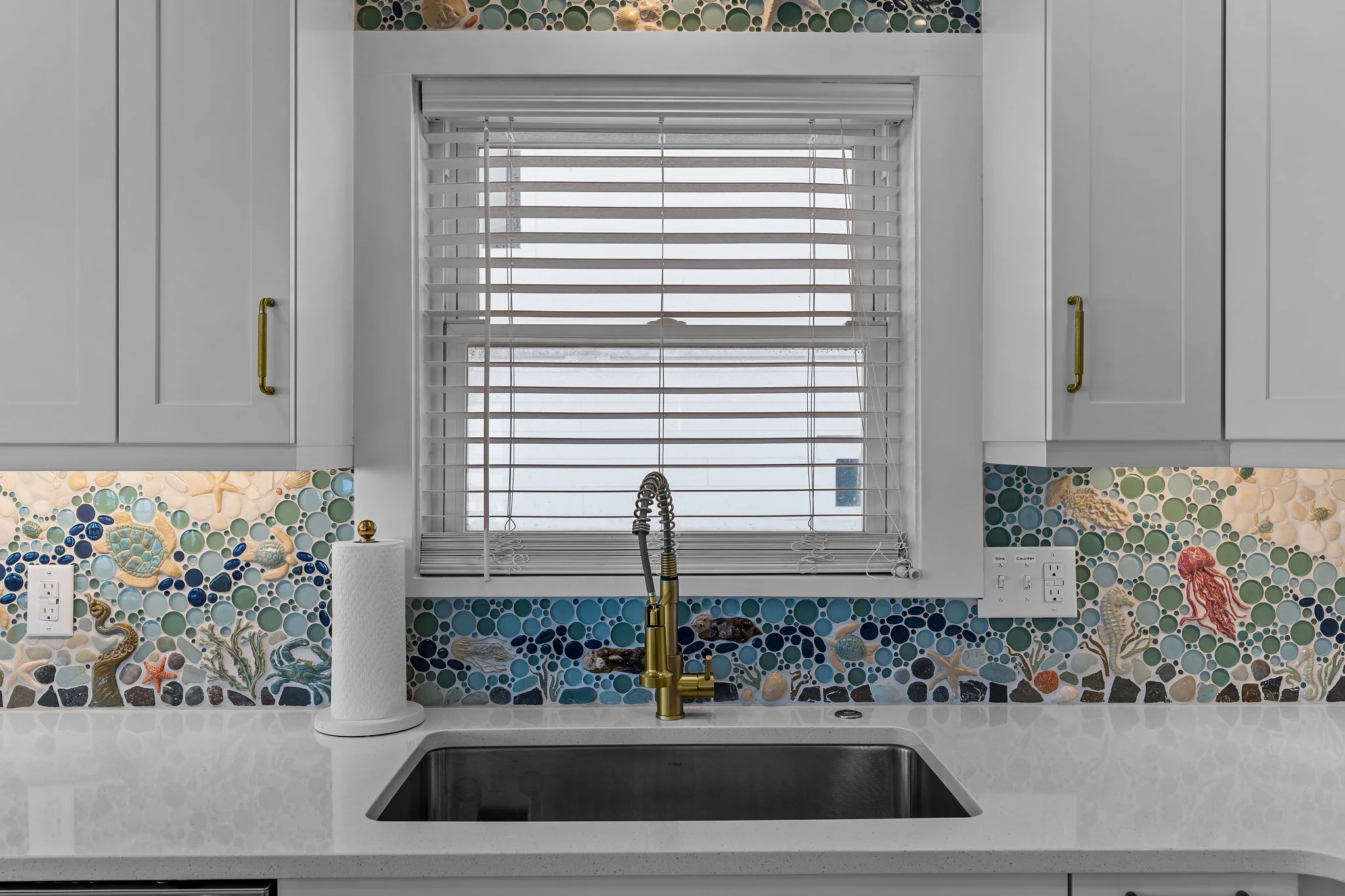 A kitchen with a window above the sink, white cabinets with gold handles, a mosaic tile backsplash featuring sea creatures and sea shells, a paper towel roll, and electrical outlets on the countertop.