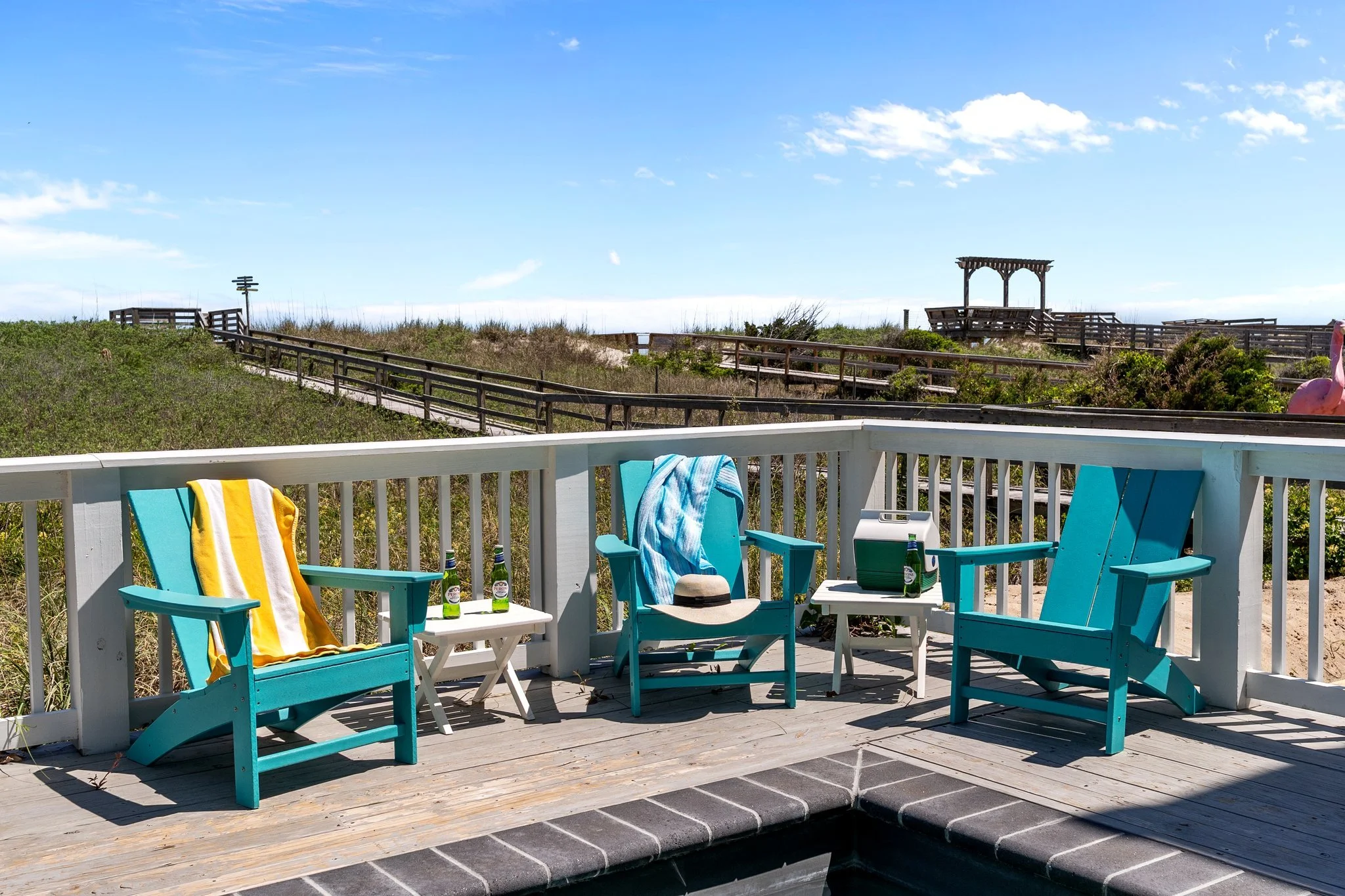 View from a porch with turquoise chairs, a small white table with drinks, towels, and a wide wooden railing. Beyond the porch are sand dunes, grassy vegetation, and a wooden walkway leading to a gazebo on a beach. The sky is blue with some white clou
