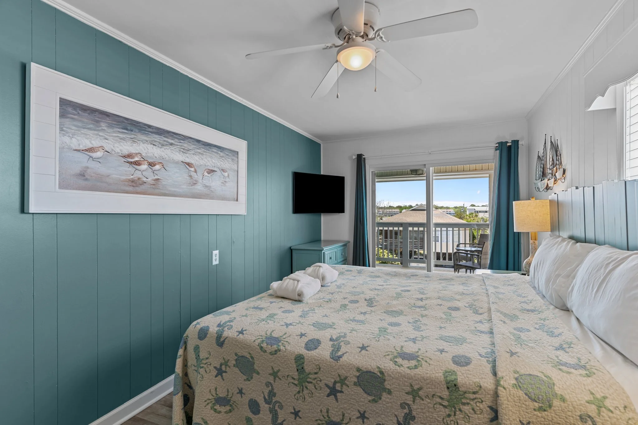 A bedroom with a teal accent wall, a large painting of beach sandpipers on the wall, a tropical-themed quilted bedspread, and a sliding glass door leading to a balcony with a view of neighboring houses and trees.