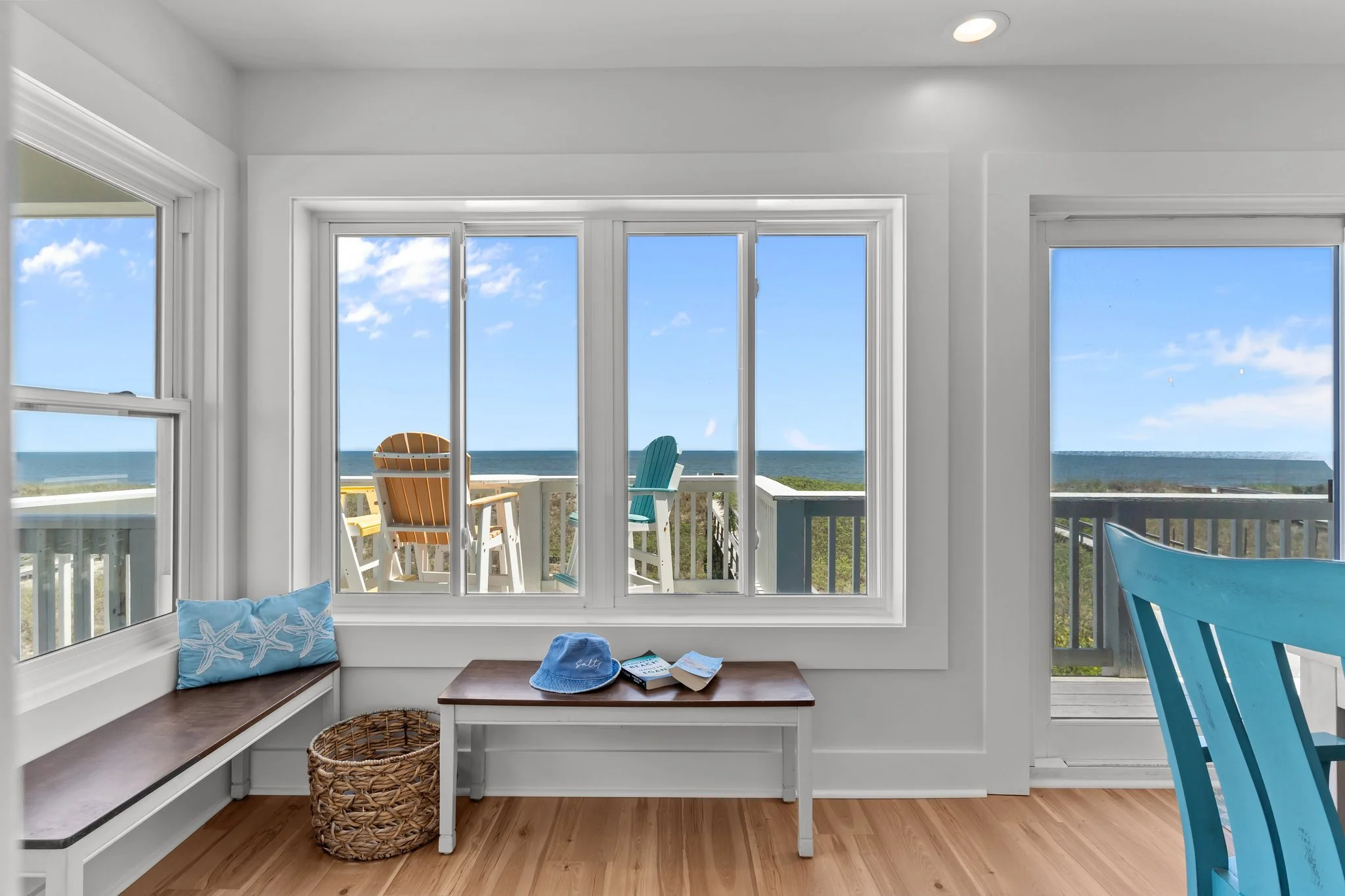Interior view of a room with large windows overlooking a beach and ocean. The room has wooden flooring, a built-in window seat with pillows, a small table with hats and magazines, and a patio area with Adirondack chairs outside.