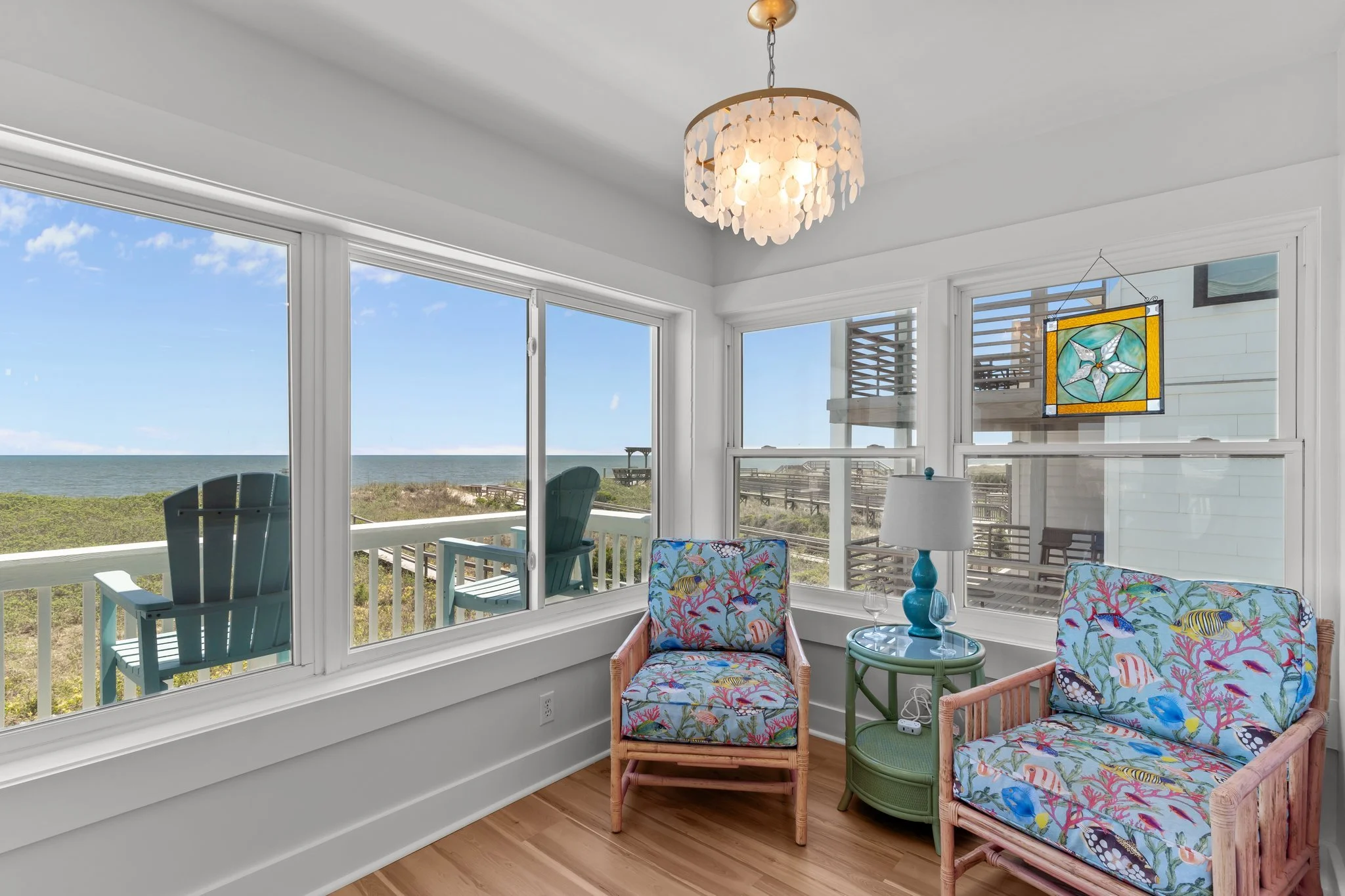 Sunroom with large windows showing ocean view, wicker chairs with colorful tropical-patterned cushions, blue lamps on side table, stained glass wall hanging, and chandelier overhead.