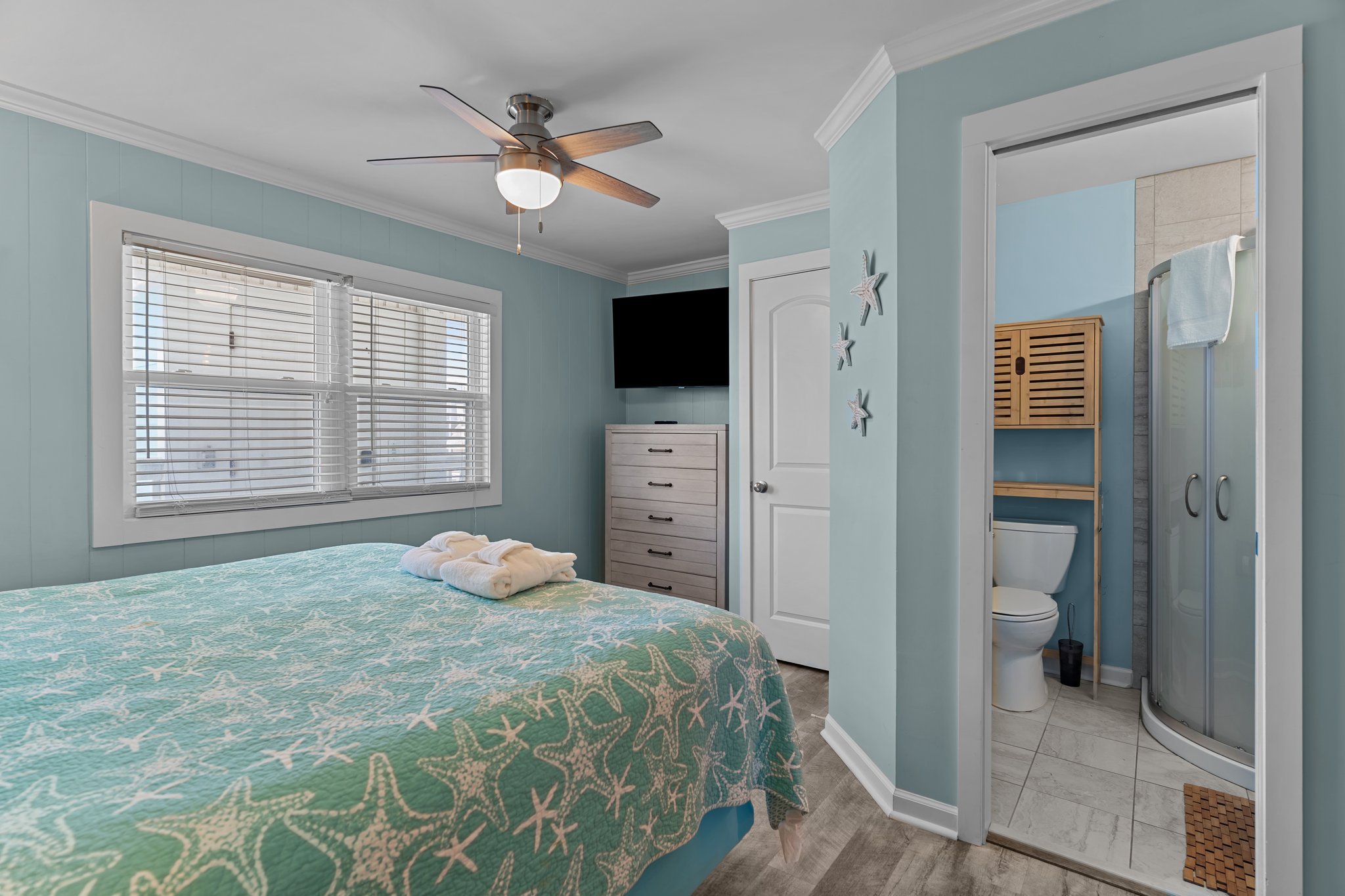 A bedroom with light blue walls, a bed with blue starfish-patterned bedding, a window with blinds, a ceiling fan with wooden blades, a flat-screen TV on the wall, a white dresser, and a view into a bathroom with a toilet, shower, and towel.