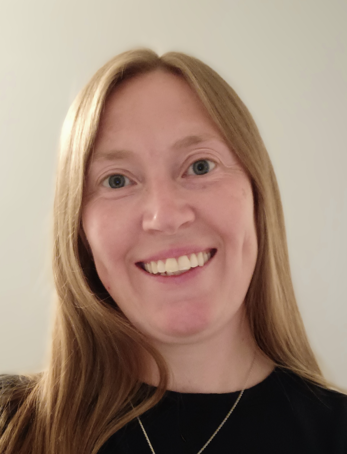 Close-up of a smiling woman with blonde hair, blue eyes, and fair skin, wearing a black top and a necklace, against a plain white background.