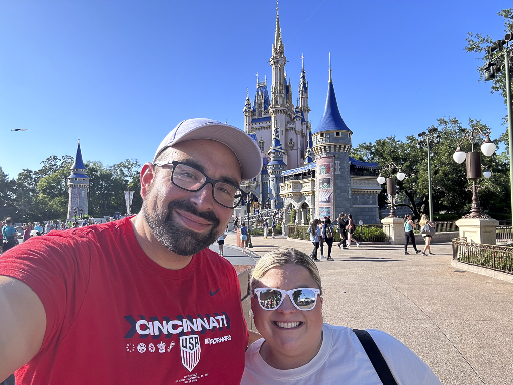 A man and woman smiling for a selfie in front of Cinderella's Castle at Disney World, with clear blue sky and park visitors in the background.