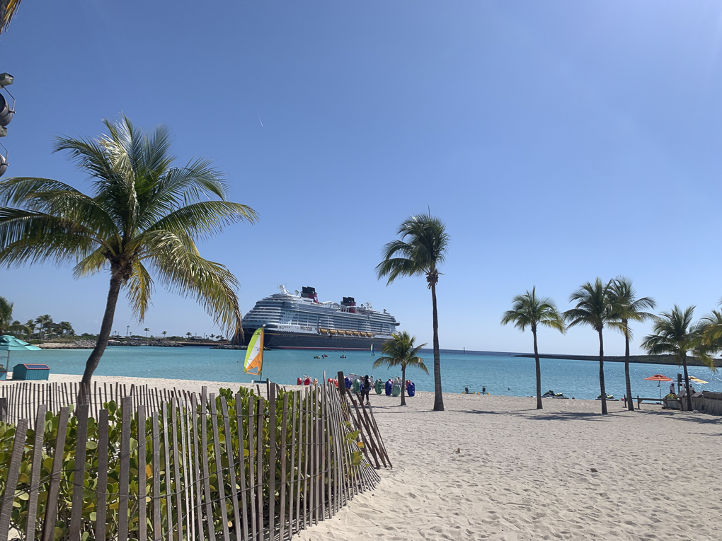 Beach scene at Castaway Cay with palm trees, sandy shore, and a Disney cruise ship in the background on the water.