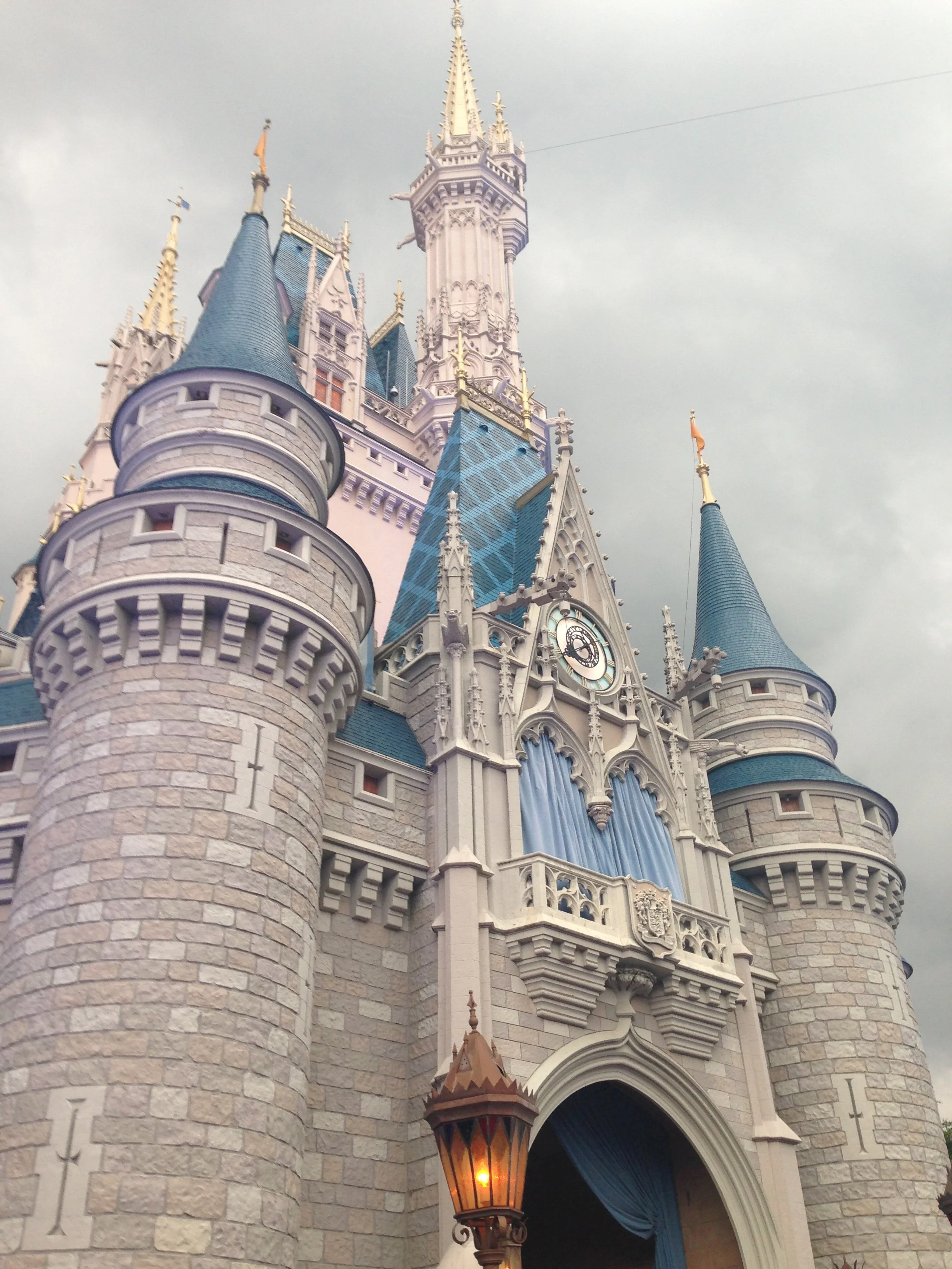 Cinderella's Castle at Disney World. Castle with stone walls and blue spires, decorated with flags and ornate details, with a lantern in front and cloudy sky in the background.