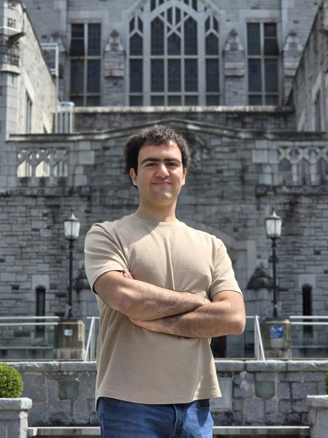A young man with crossed arms standing in front of a historic stone building with large windows and decorative architecture.