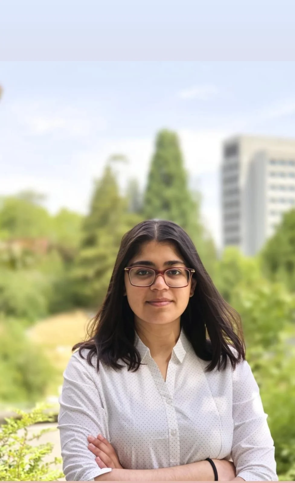 Prajna, a young woman with dark hair, glasses, and a white shirt standing outdoors with her arms crossed, trees, and buildings in the background.