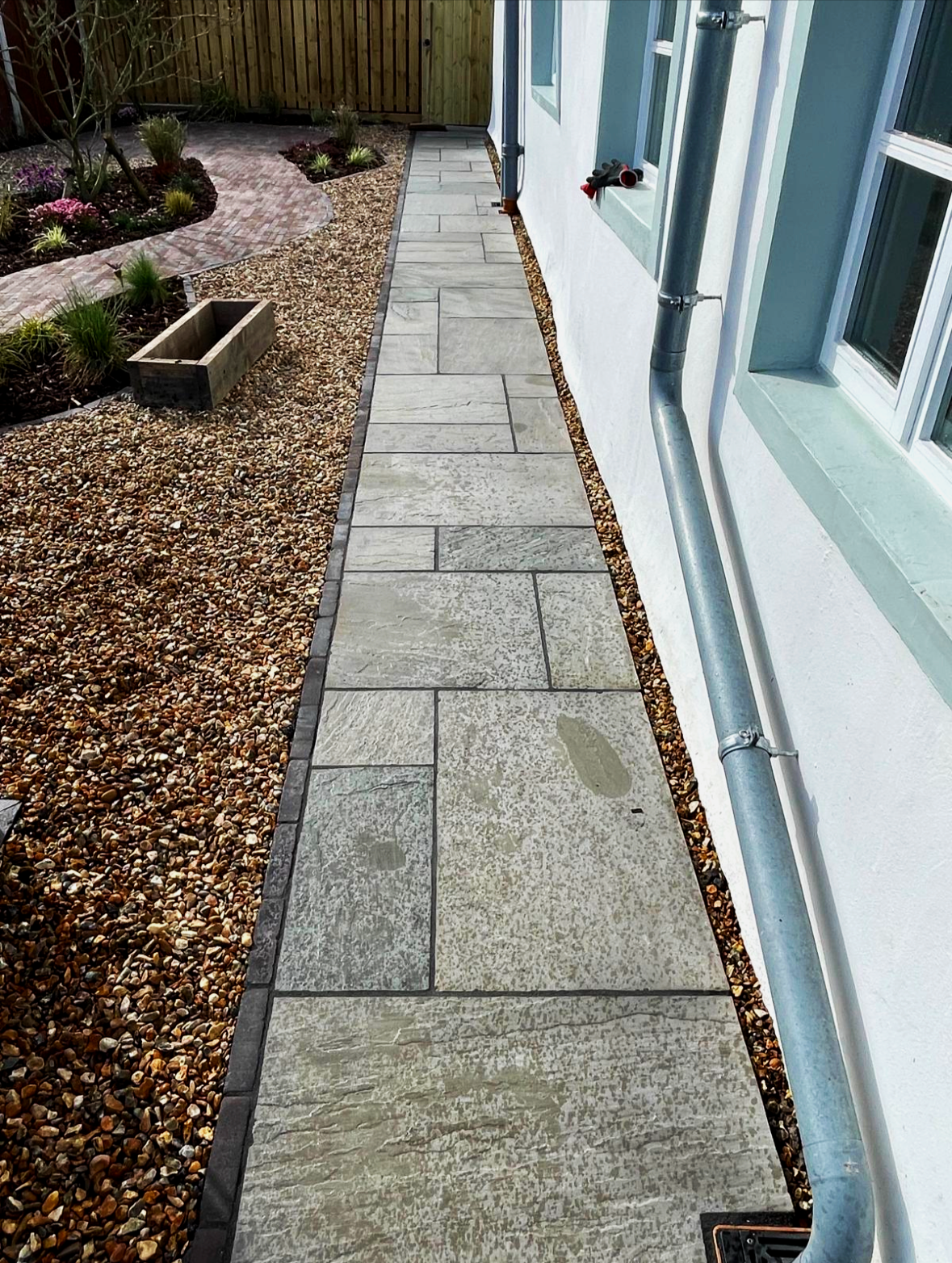 A newly installed concrete patio running alongside a white house wall, with a garden on one side and a drainpipe attached to the house.