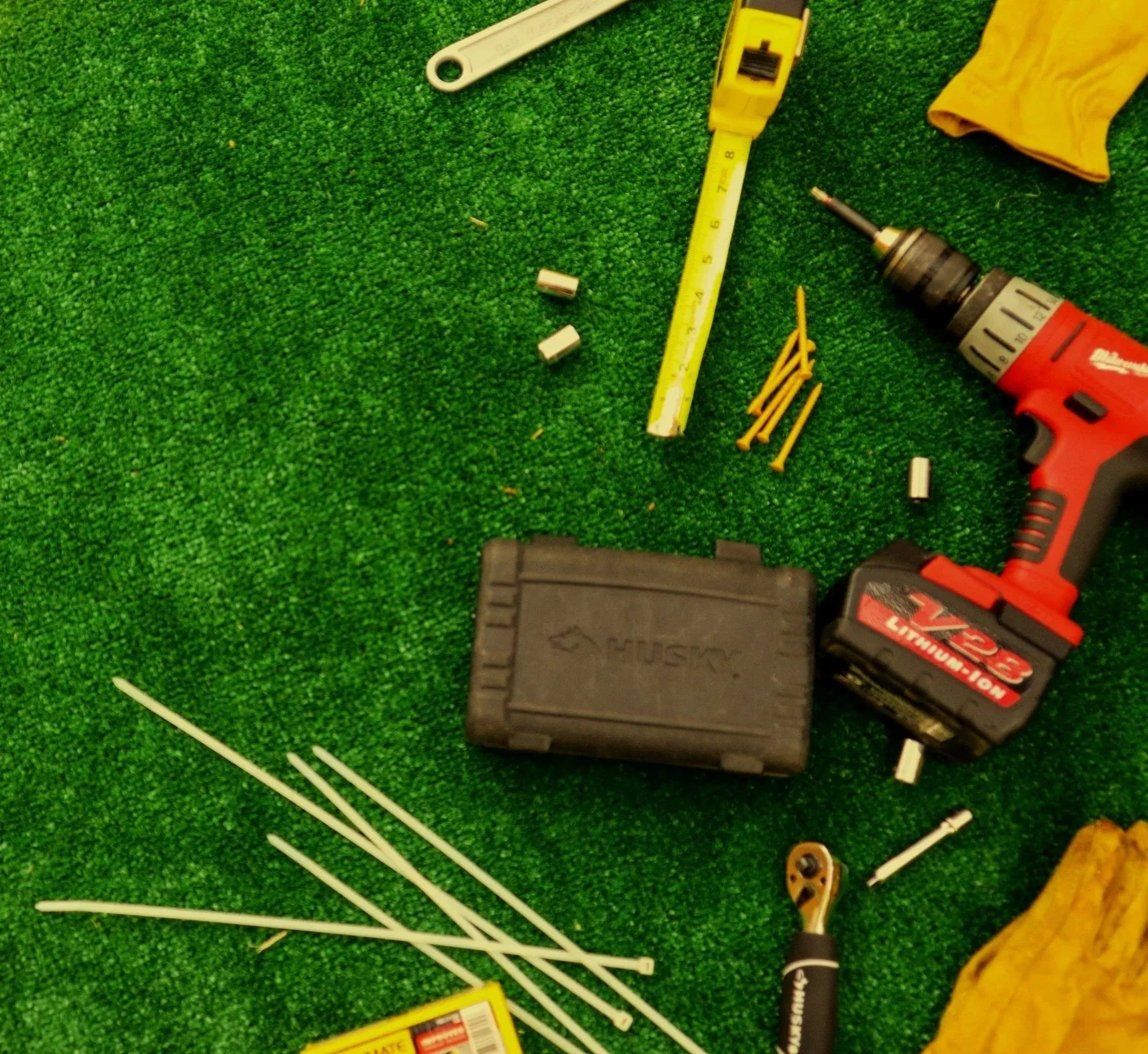 Tools and materials on green artificial grass, including a yellow measuring tape, a red cordless drill, a black toolbox, yellow zip ties, and white sticks.