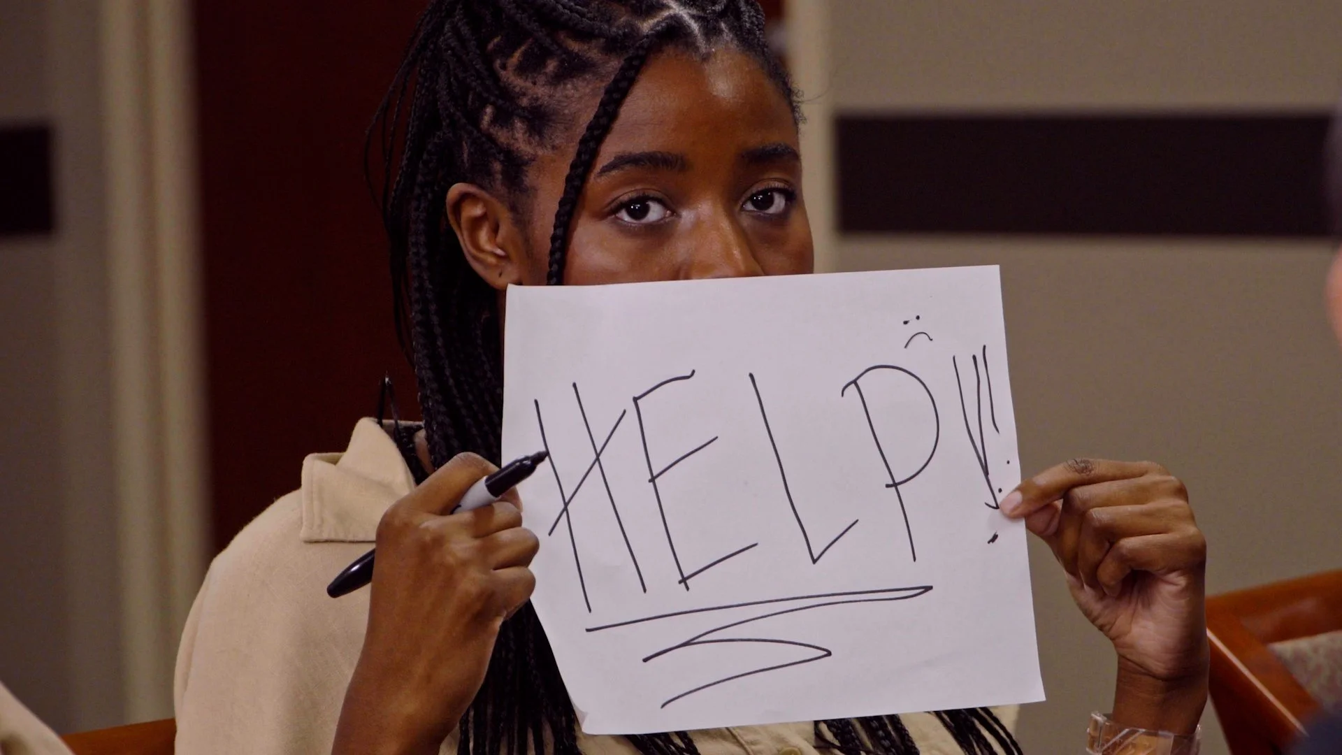 A woman with braided hair holding a sign that says "HELP!" with a sad face drawn on it, in an indoor setting.
