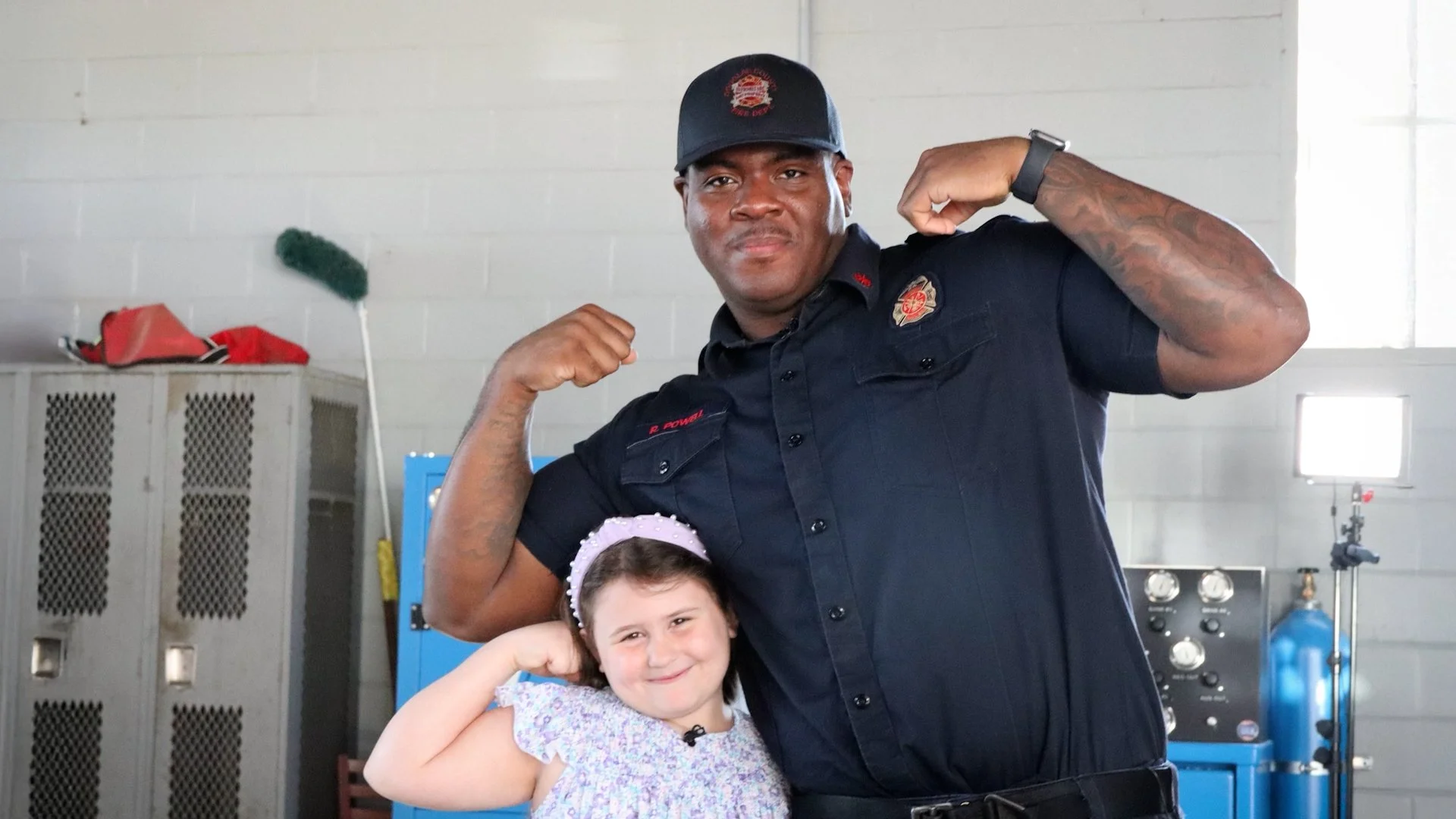 A firefighter and a young girl are posing together, both flexing their arms in a display of strength inside a fire station.