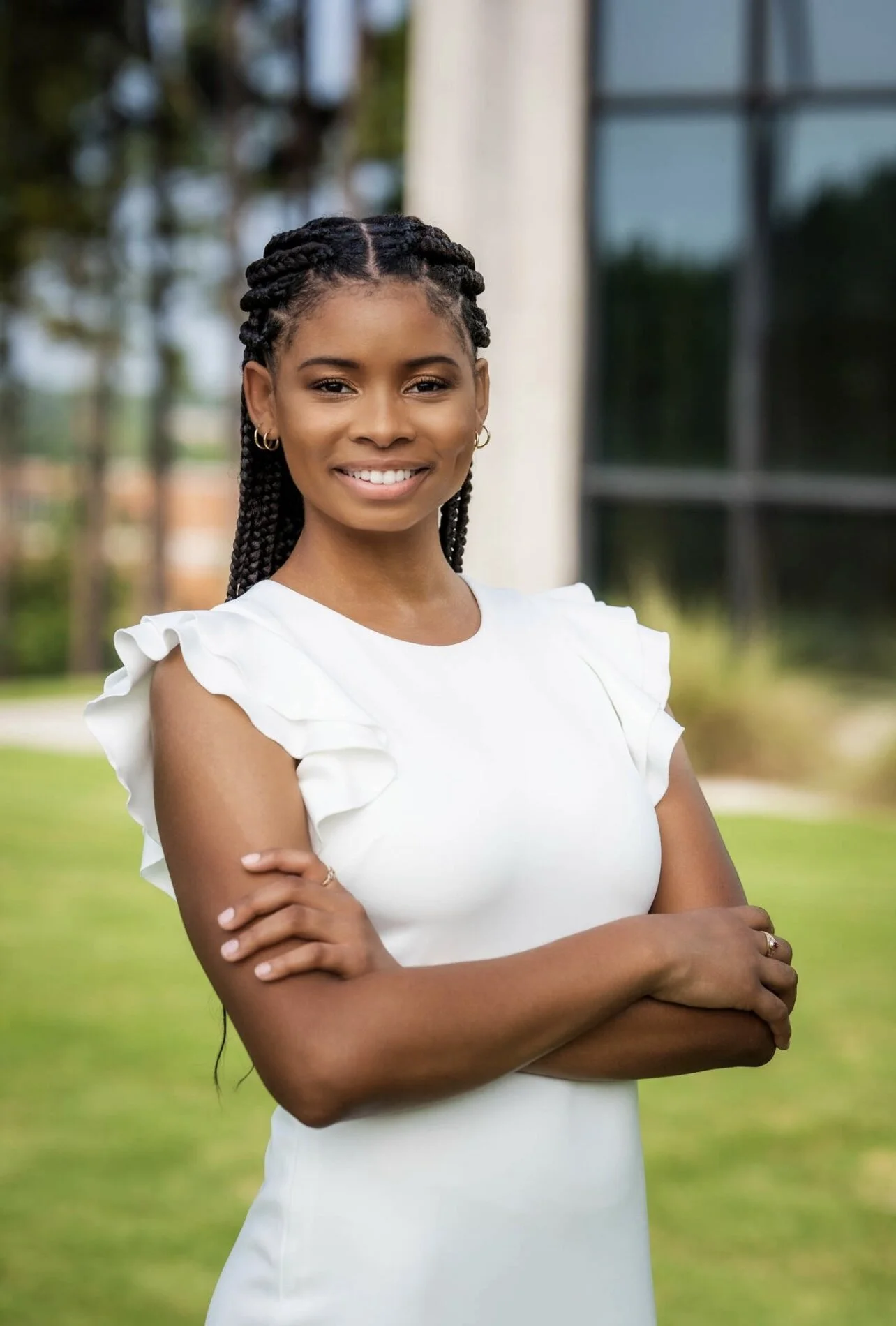 Young woman with braided hair smiling, standing outdoors in front of a building and greenery.