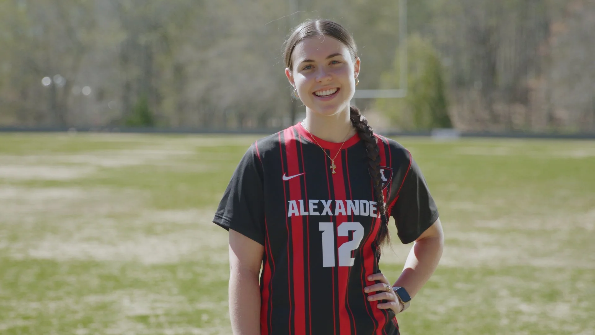 Young woman with braided hair smiling on a sports field, wearing a black and red soccer jersey with 'ALEXANDER' and the number 12.