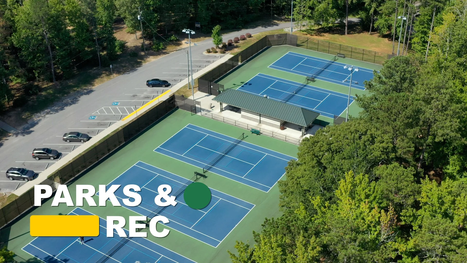 An aerial view of a sports complex with multiple tennis courts, some shaded by trees and others open, surrounded by a parking lot with a few cars, and lush greenery in the background.