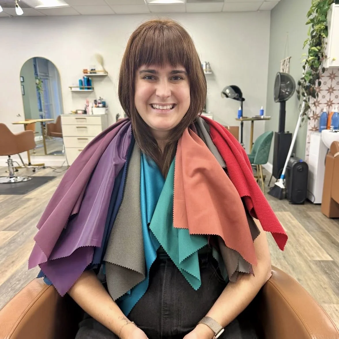 Woman sitting in a salon with hair samples draped over her shoulders, smiling at the camera.