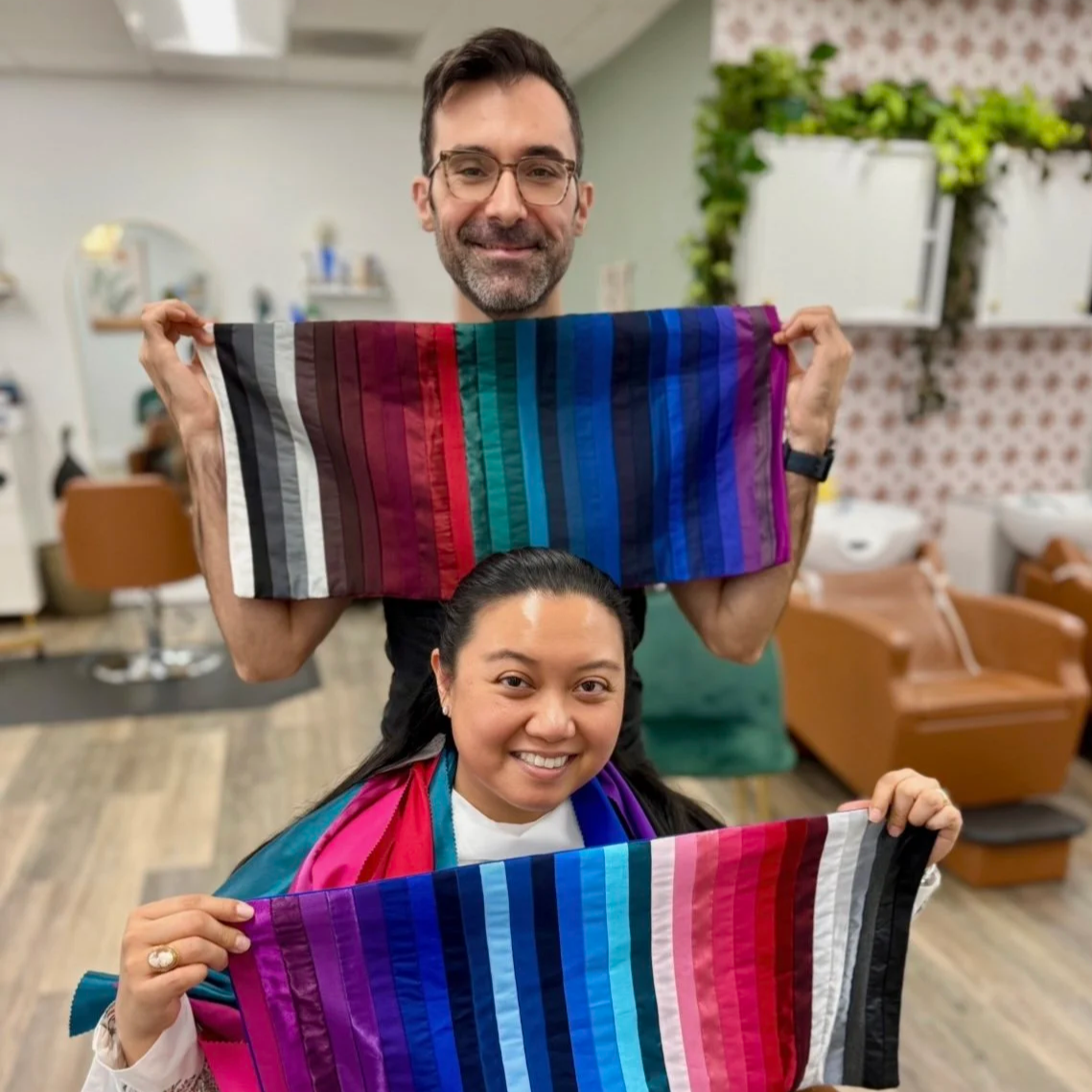 A man and a woman smiling, each holding a colorful striped fabric, with a salon or barbershop background.