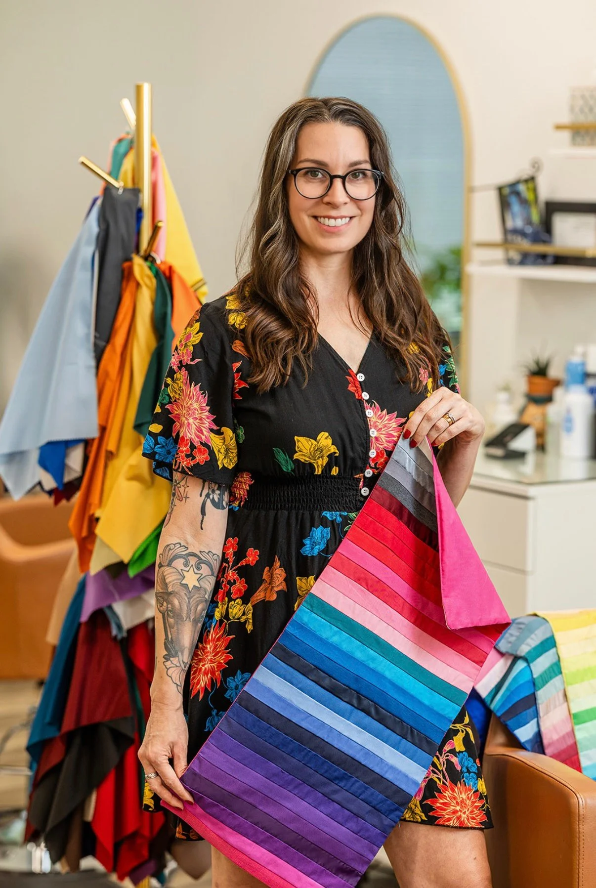A smiling woman with glasses and long brown hair holding a rainbow-colored fabric at a fabric store, with colorful fabric bolts on display behind her.