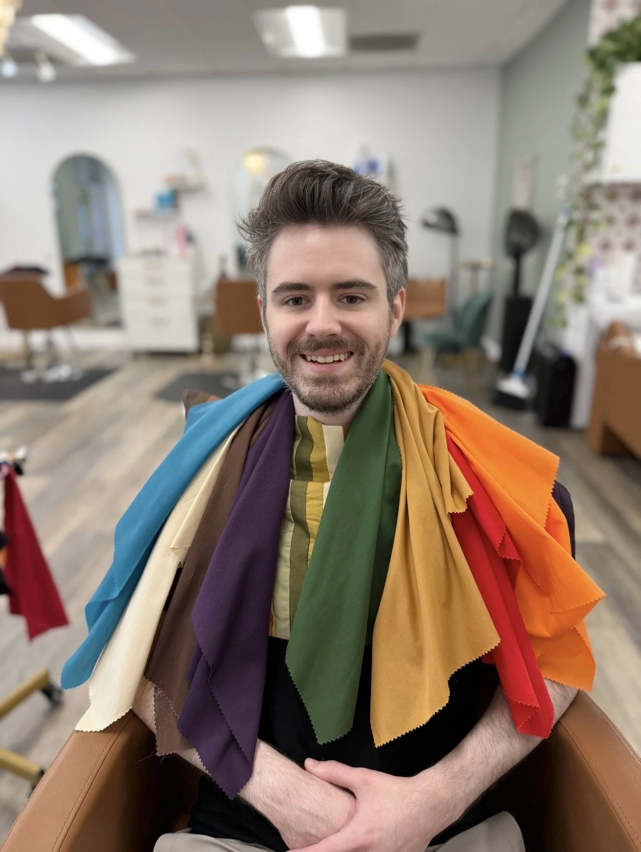 A man sitting in a salon chair, smiling, with a rainbow-colored fabric draped over his shoulders, in a hair salon with chairs and mirrors in the background.