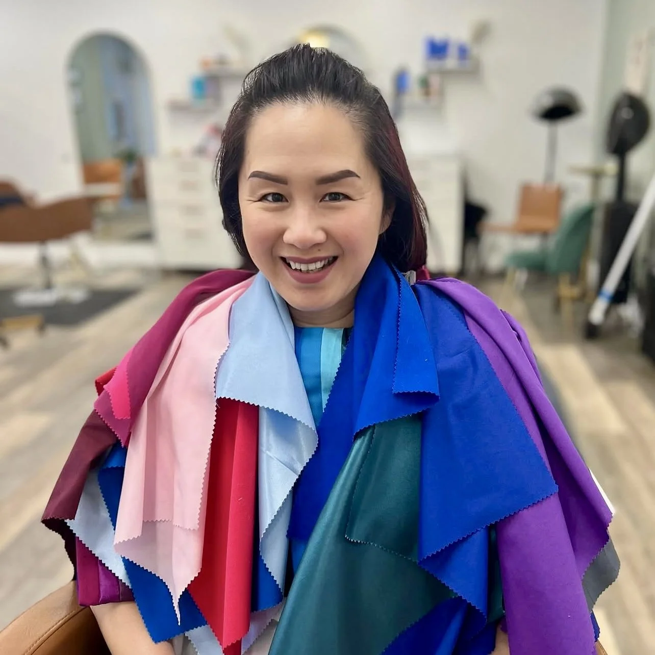 Woman smiling with hair up, wearing a colorful cape made of fabric swatches in pink, red, light blue, dark blue, purple, and teal, in a salon.