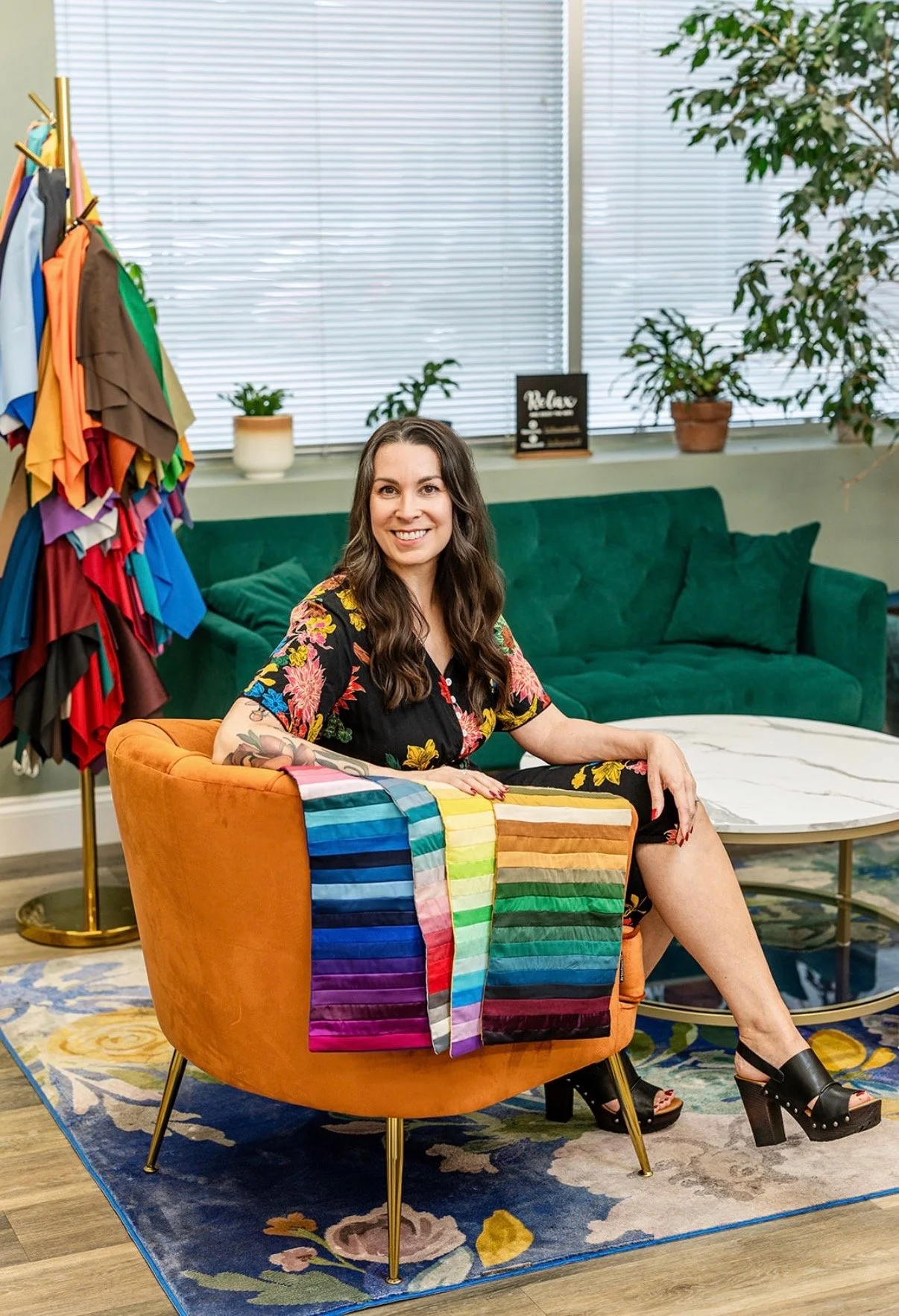 A woman with long brown hair wearing a floral dress is sitting on an orange armchair. She is smiling and has rainbow-colored fabric draped over the armrest. Behind her, there is a green sofa, a round white coffee table, and a dressing rack with colorful fabric. The room has large windows with blinds, potted plants, and decorative signs on the windowsill.