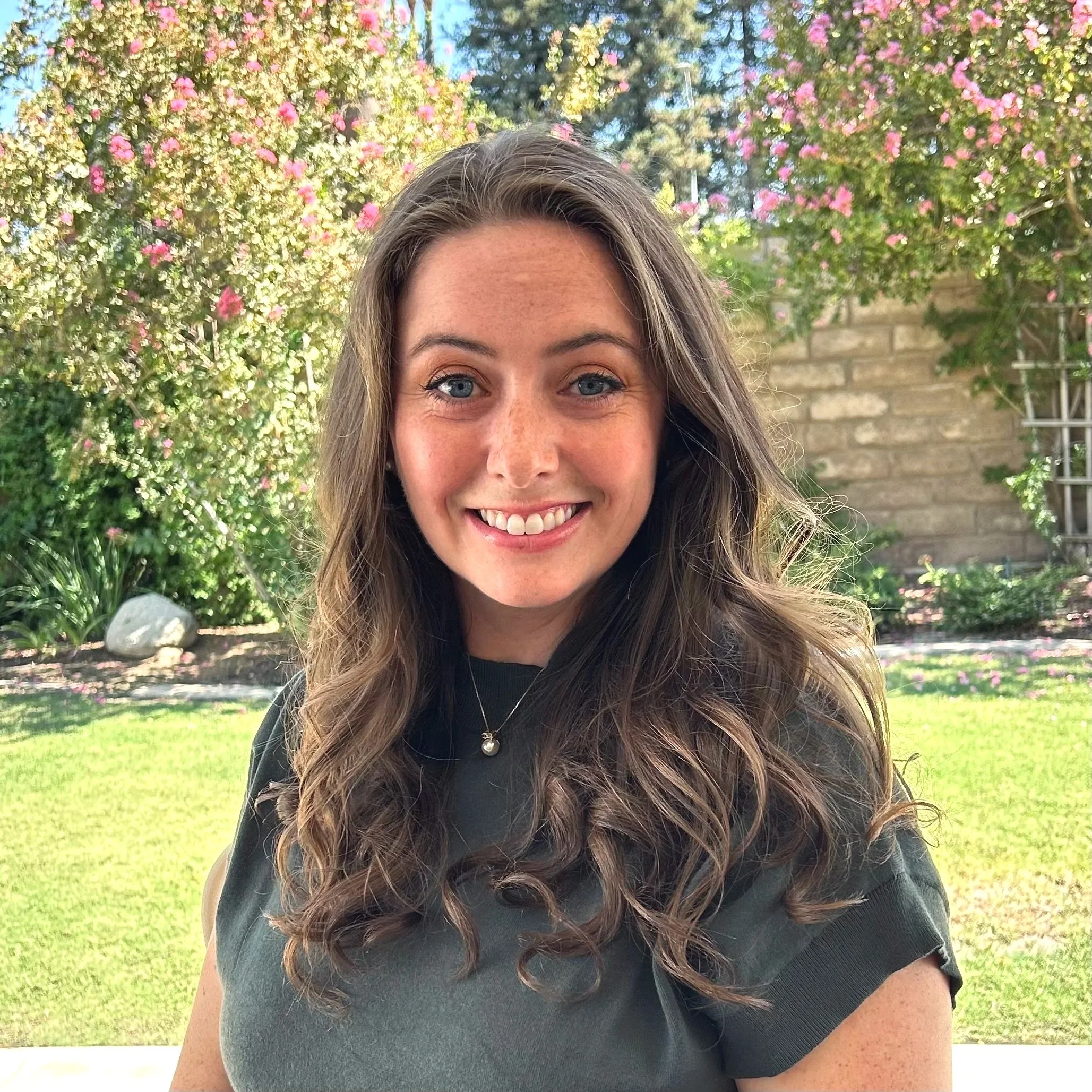 A woman smiling outdoors with long, wavy brown hair, wearing a black top, standing in front of flowering bushes with pink flowers and trees in the background.