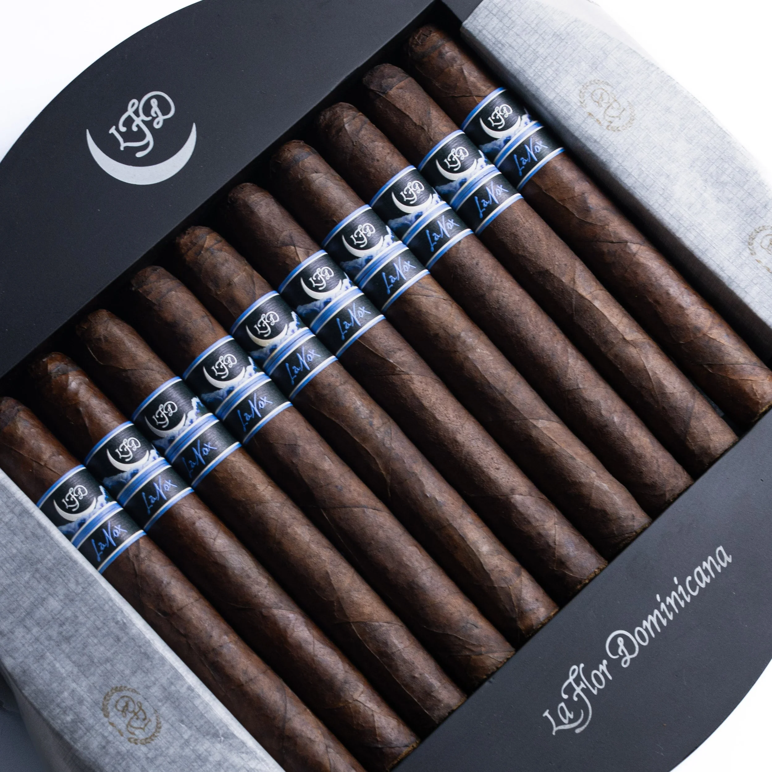 A black box containing five cigars with dark brown wrappers and blue and black bands with white and blue text. The box has a silver logo and decorative parchment paper with gold accents.