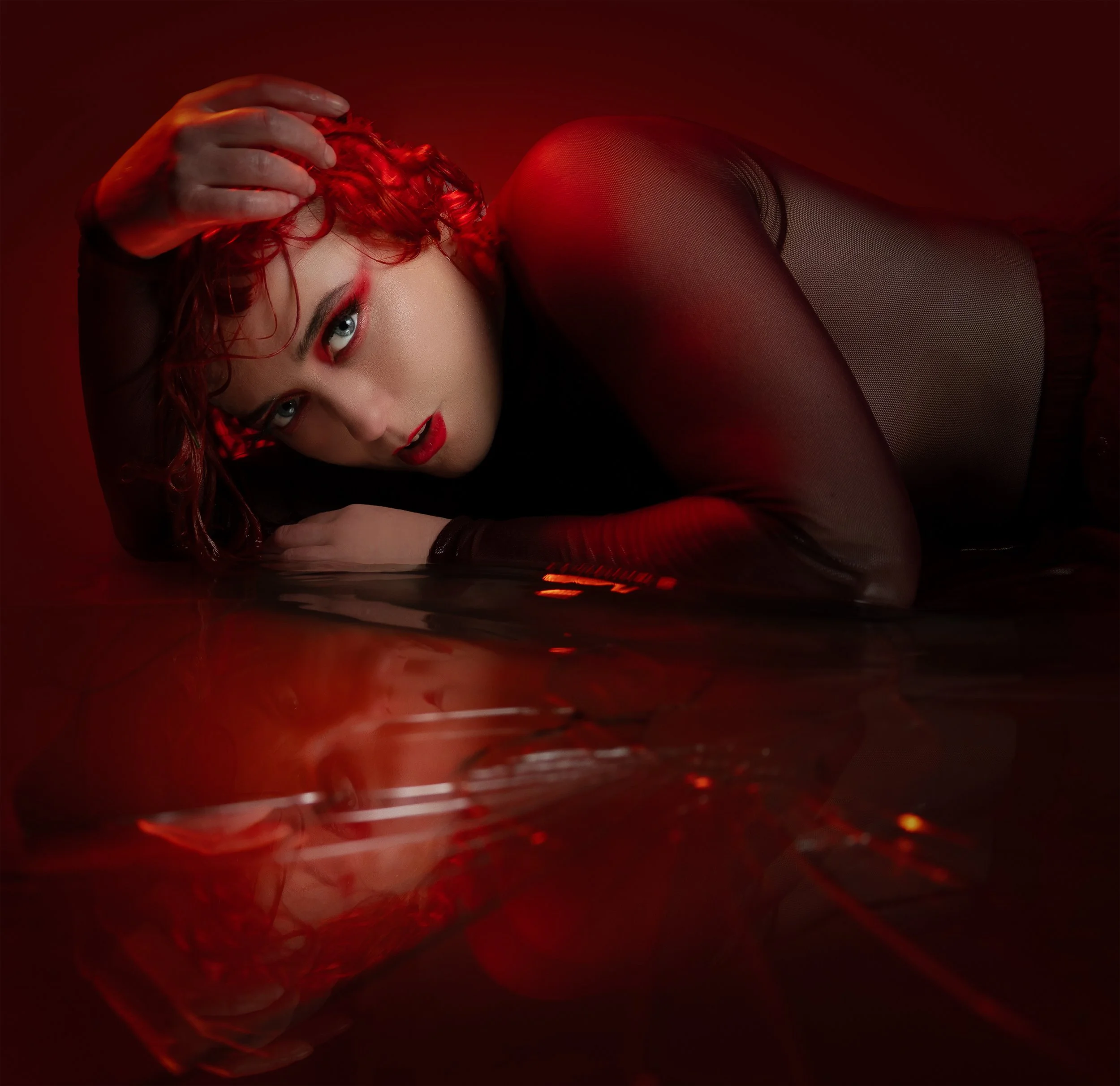 A woman with curly red hair lying on a reflective surface, illuminated by red lighting, wearing fishnet stockings and dark clothing, with eye makeup and red lipstick.