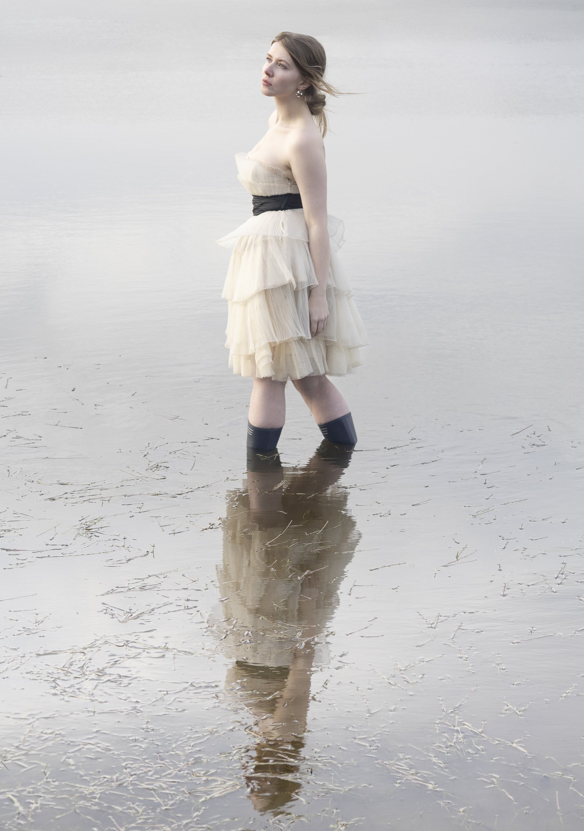 A young woman with light skin and long brown hair standing in shallow water at the edge of a lake or pond. She is wearing a strapless cream-colored dress with ruffles, a black waistband, and knee-high black boots. Her hair is slightly windswept, and 