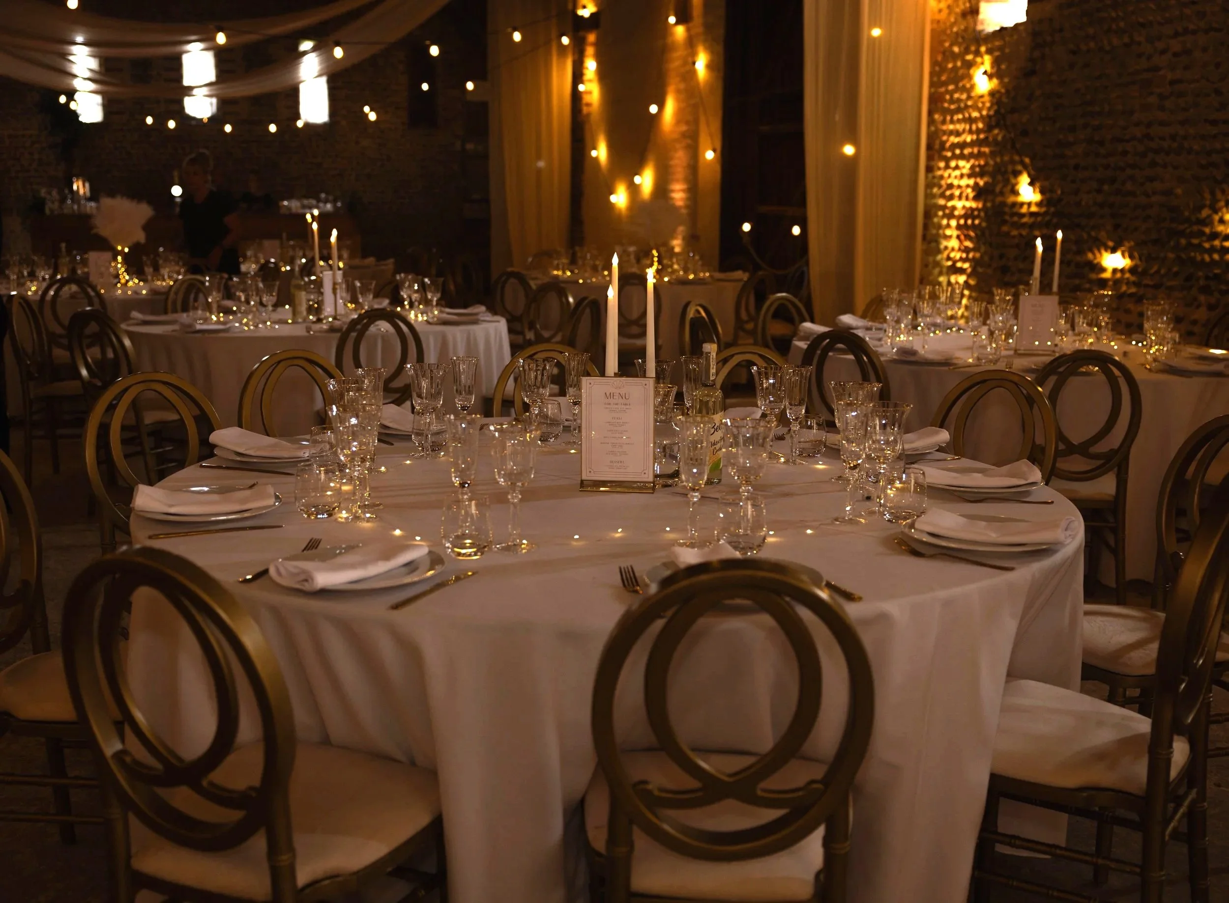 A dimly lit banquet hall decorated for an event with round tables covered in white tablecloths, each set with glassware, silverware, napkins, and a menu card. Tall candles are on the tables, and warm string lights and a textured brick wall with sheer