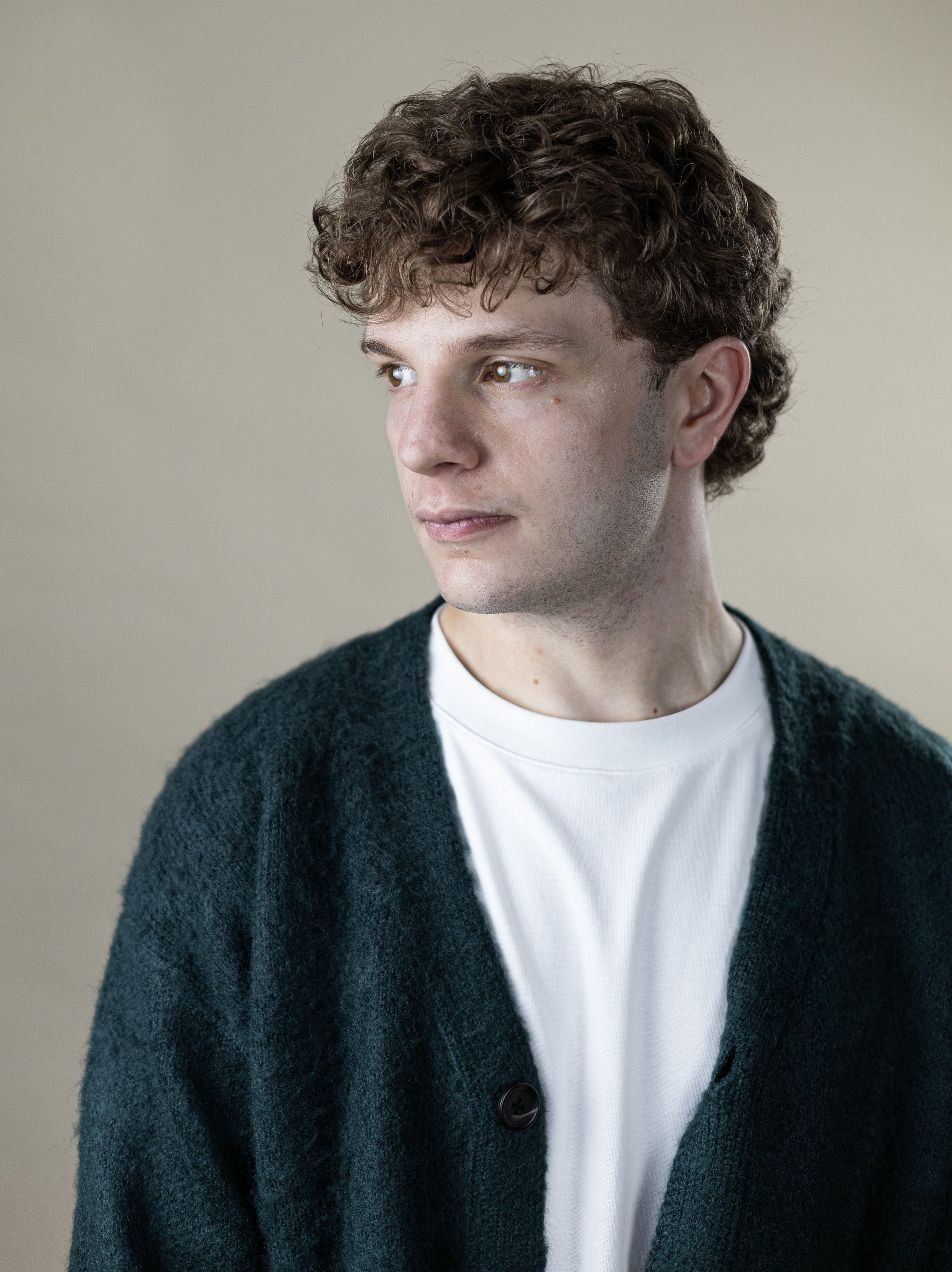 Close-up of a young man with curly brown hair wearing a white T-shirt and a dark green cardigan, looking to his left, against a neutral background.