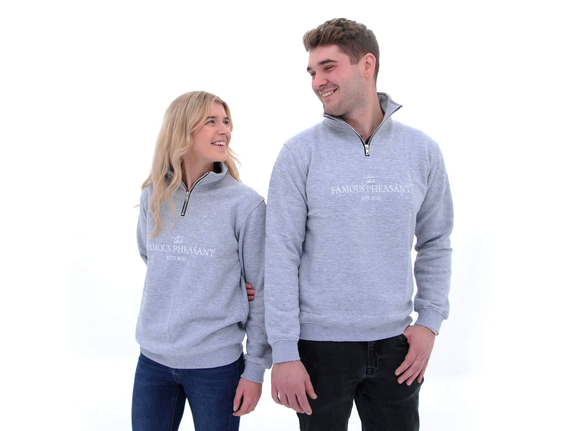 A young man and woman wearing matching gray sweatshirts with the text 'the FAMOUS PHEASANT ESTD 2021' on the front, smiling and looking at each other against a plain white background.