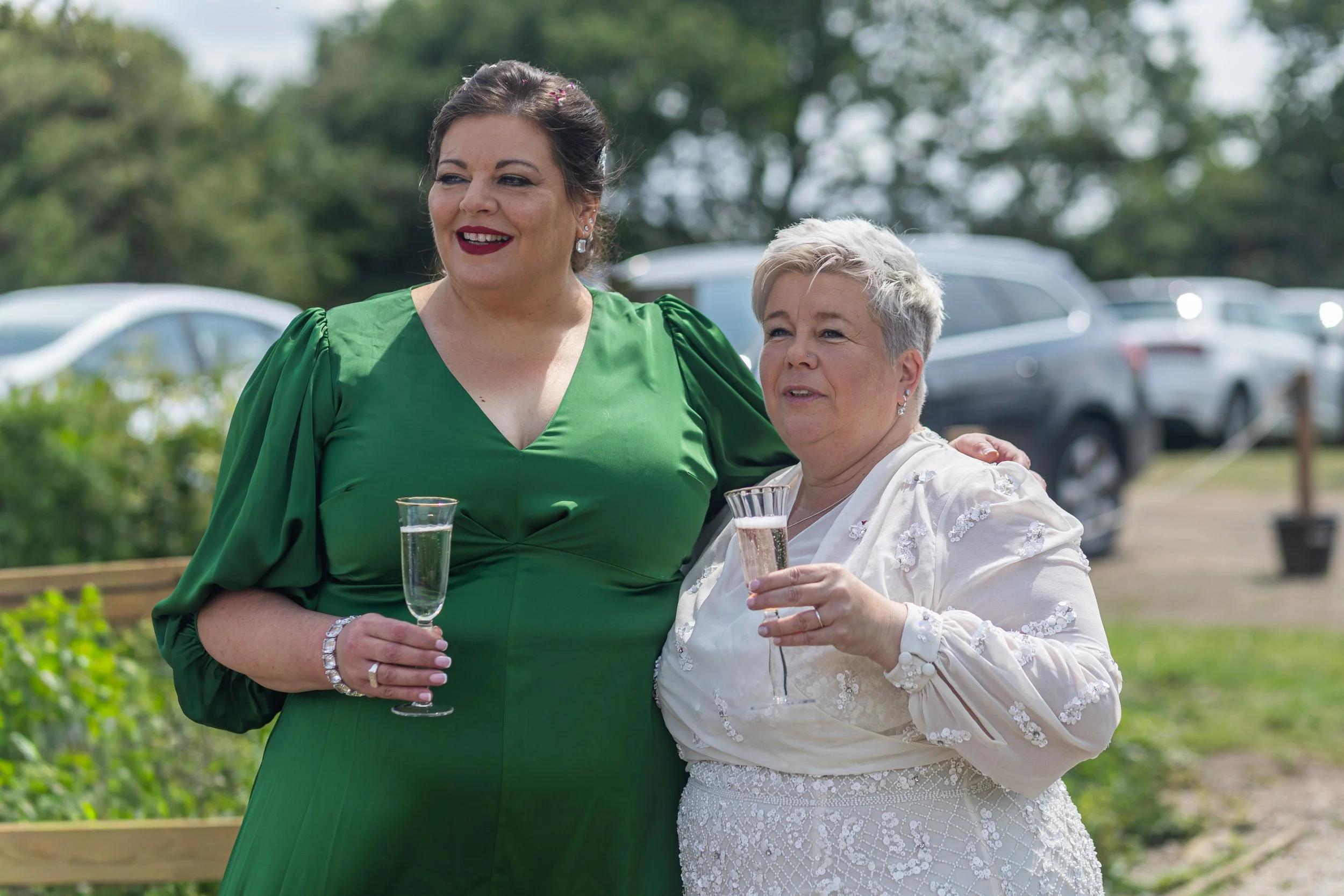 Two women standing outdoors, holding glasses of champagne, smiling, with a background of trees and parked cars, dressed in formal attire.