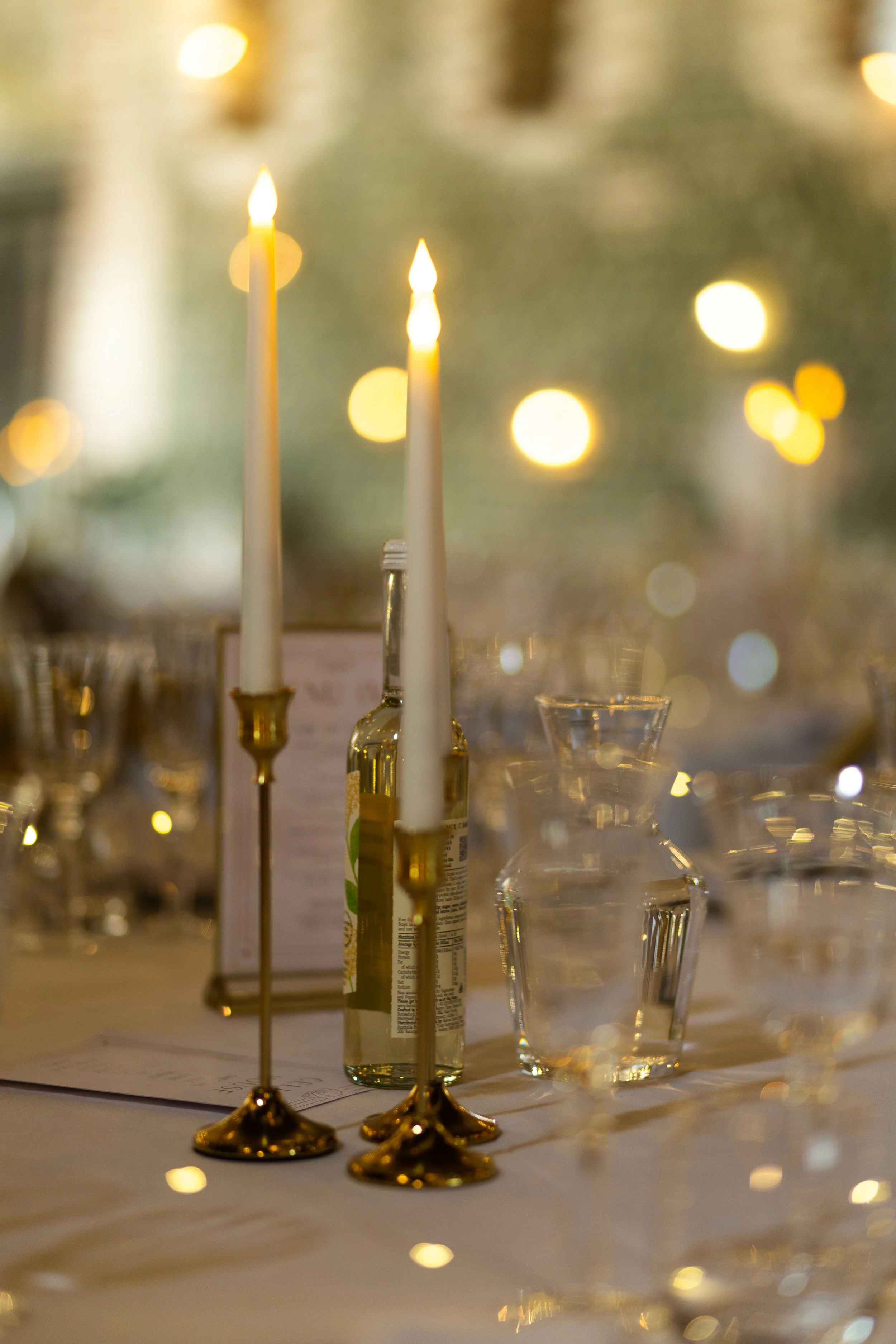 Two white candles in gold holders, surrounded by glassware and blurred warm lights in the background, creating a festive or romantic atmosphere.