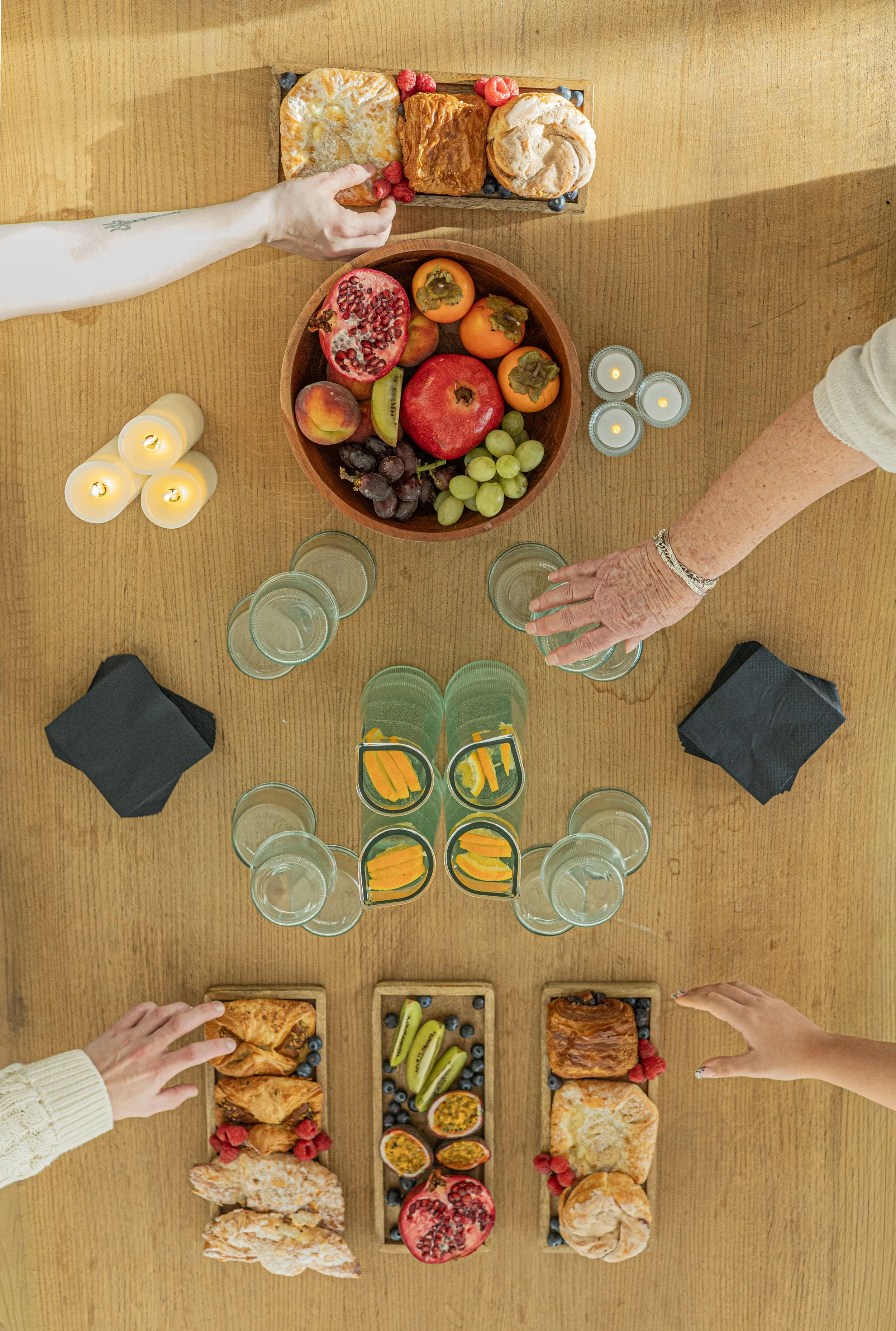Overhead view of a table with assorted foods including a large bowl of mixed fruit, pastries, small plates of sliced kiwi, blueberries, raspberries, and peaches, along with glasses of water, candles, and napkins.