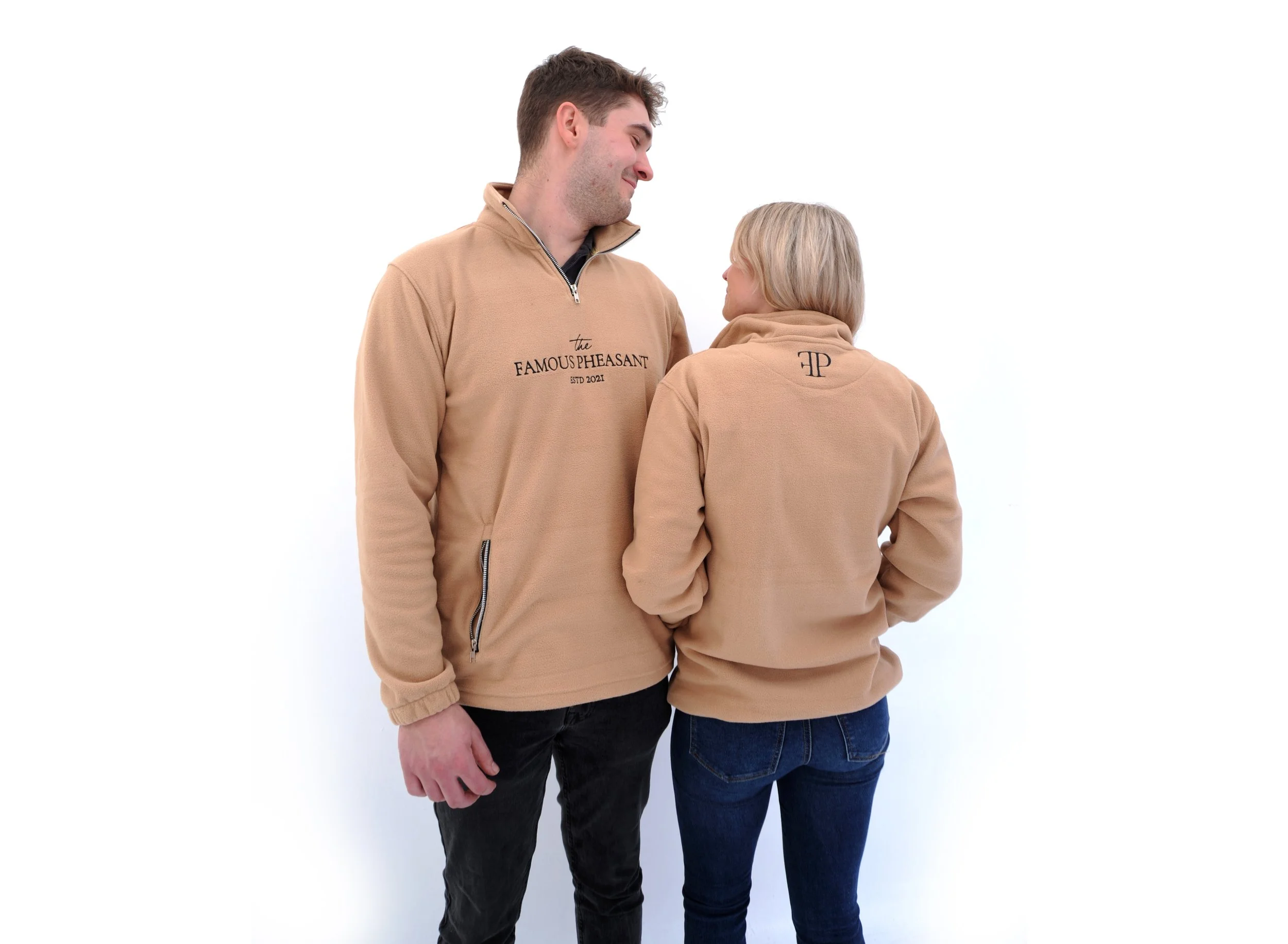 A young man and woman standing close together, both wearing matching beige sweatshirts. The man is smiling and looking at the woman, who has her back to the camera.