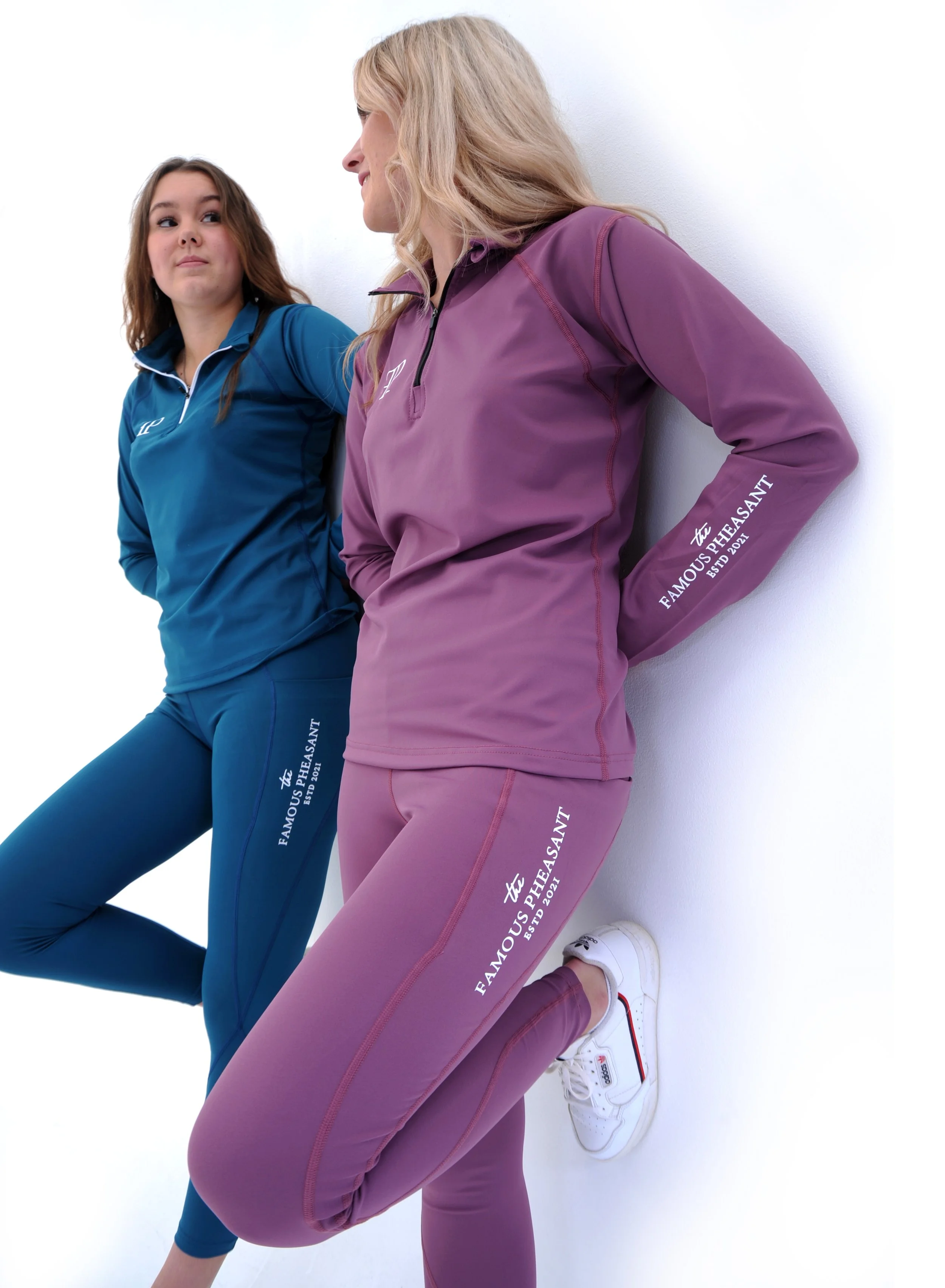 Two women in colorful athletic outfits are leaning against a white wall, engaging in a stretching or exercise routine. The woman on the left wears a blue athletic top and matching pants, while the woman on the right wears a purple athletic top and ma