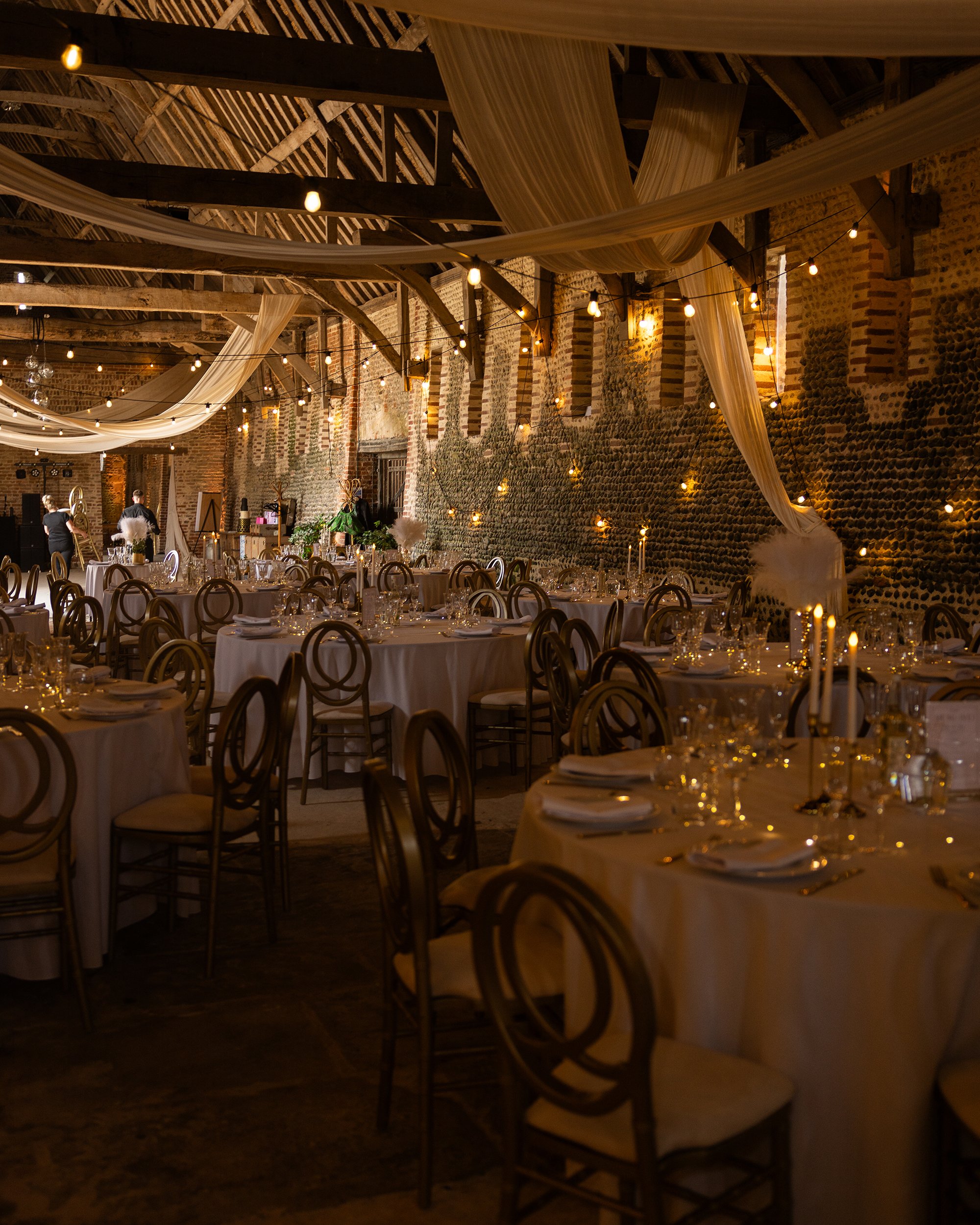 An indoor event space with round tables covered with white tablecloths, set with glassware and candles. The ceiling has draped fabric and string lights. The walls are made of exposed brick and stone. There are decorations and people preparing for an 