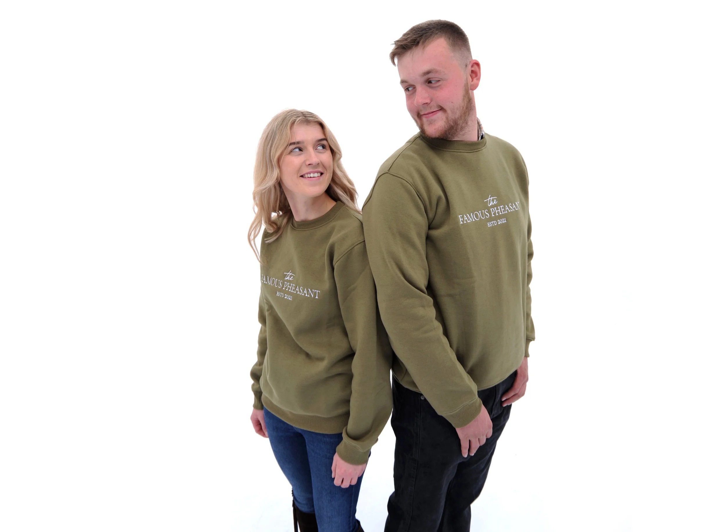 A man and woman are standing back-to-back against a white background, smiling and looking at each other. They are both wearing green sweatshirts with the text 'the FAMOUS PHEASANT ESTD 2021' on the front.