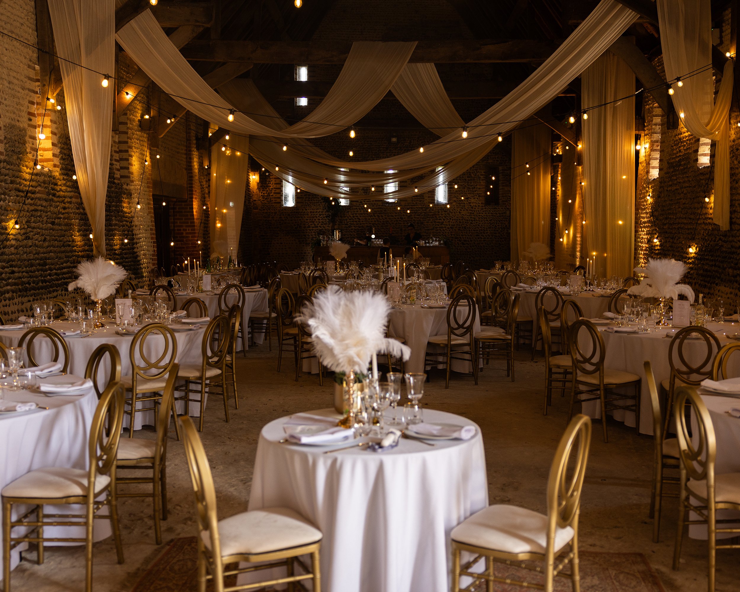Decorated banquet hall with round tables covered with white tablecloths, gold chairs, and floral centerpieces, illuminated by string lights and draped fabric on the ceiling.