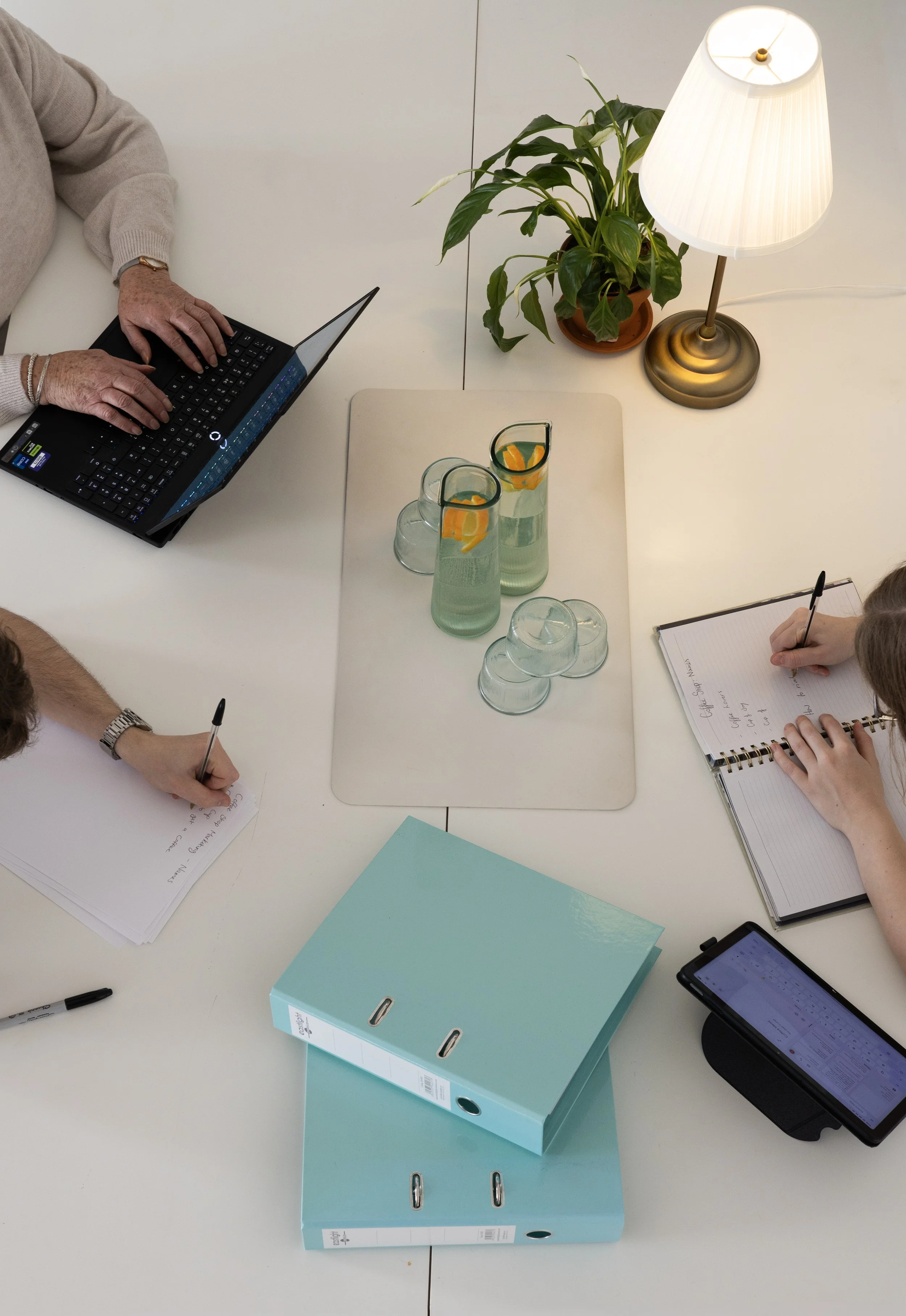 A white table with two aqua blue storage binders, a green binder, a smartphone, two glasses of water with lemon slices, a potted plant, a lit table lamp, and people writing notes.