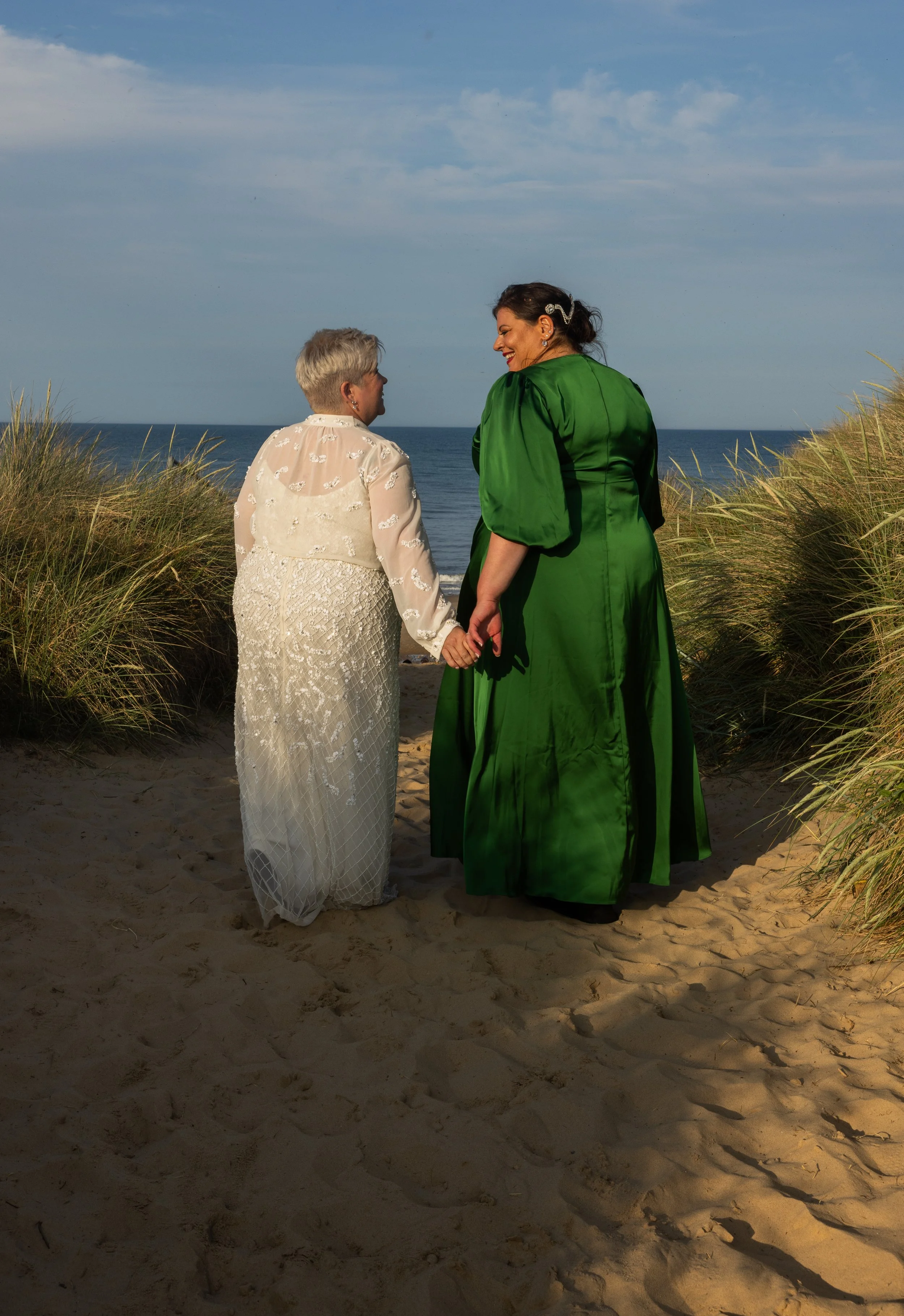 Two women holding hands on a sandy beach, dressed in evening attire, with the ocean and blue sky in the background.