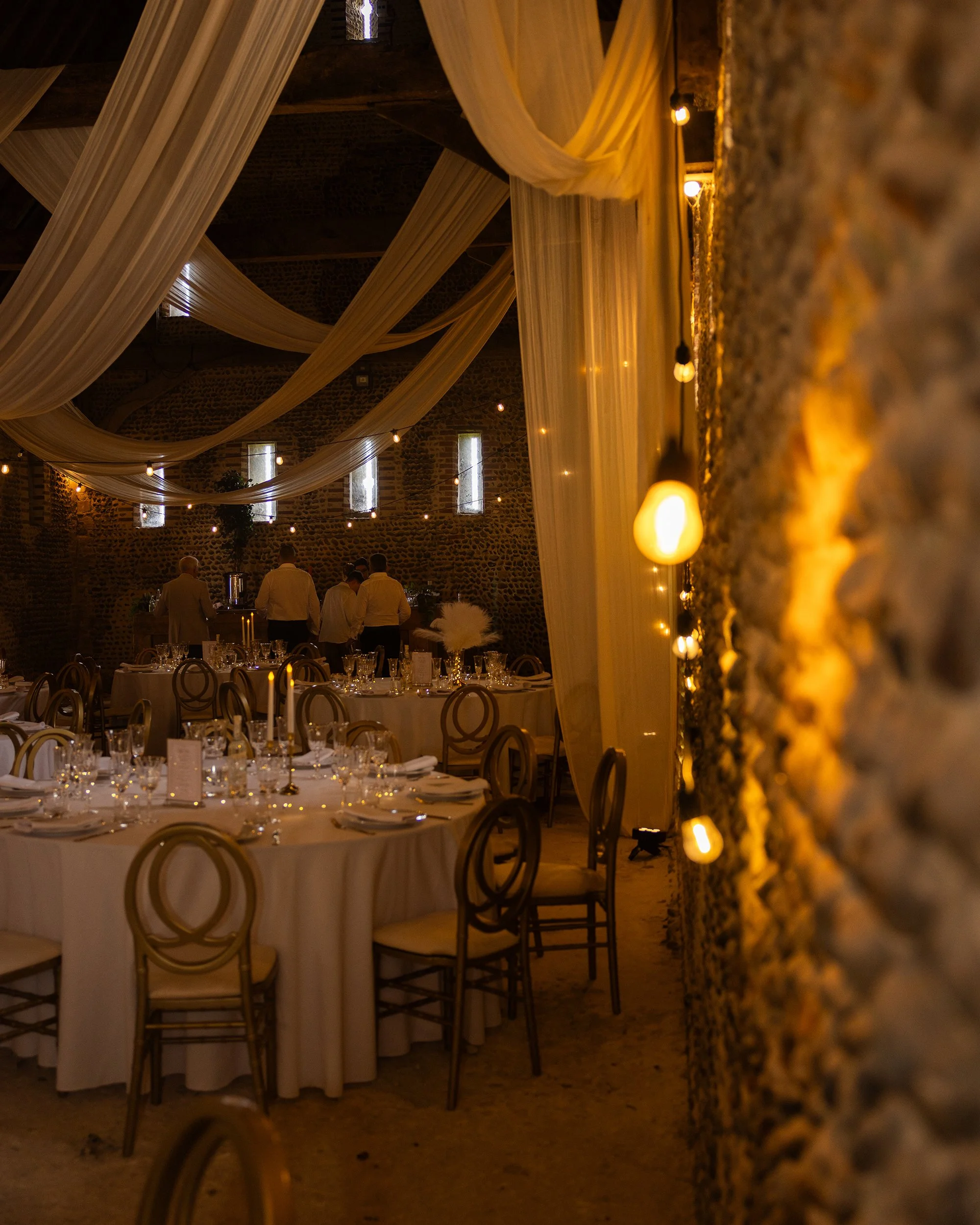 Interior of a rustic wedding or event venue with long tables set with glassware and napkins, draped fabric decoration hanging from the ceiling, warm yellow lighting, and a stone wall background.