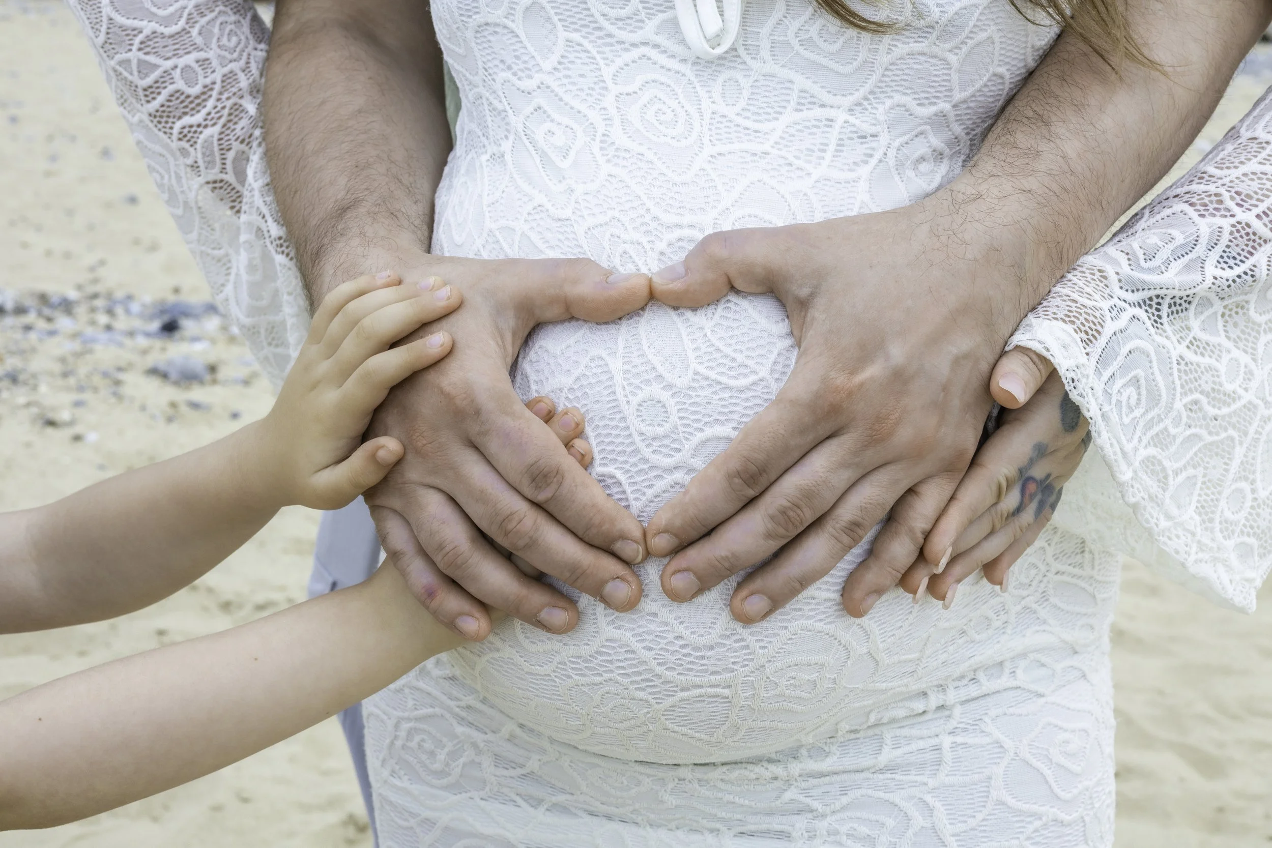 People forming a heart shape on a woman's pregnant belly, with a child's hand also included in the heart shape. They are wearing white lace clothing, and the setting appears to be outdoors on a beach.