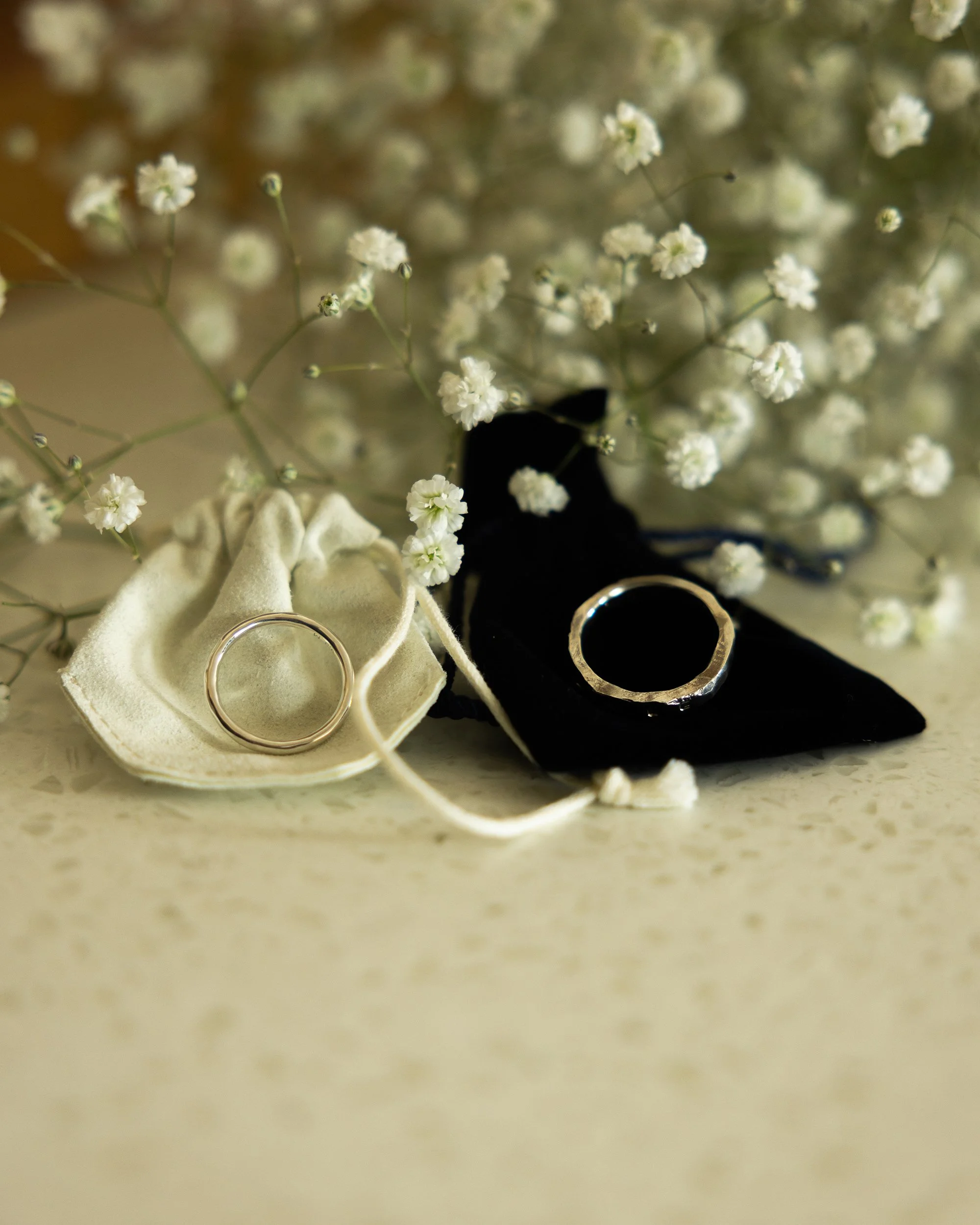Close-up of two wedding rings, one silver and one gold, resting on a soft cloth. In the background, there are white baby's breath flowers.