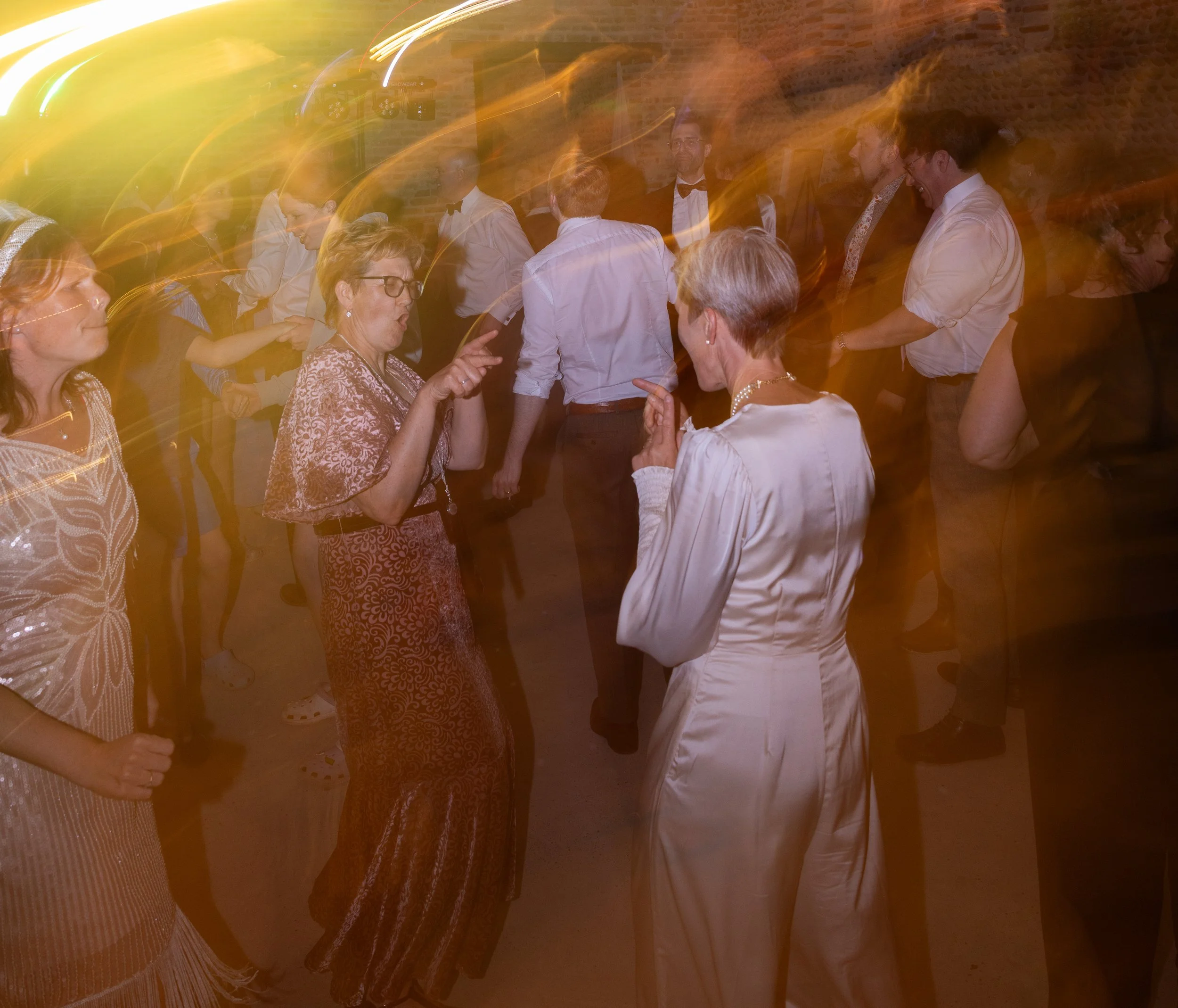 Group of people dancing and socializing at a party or celebration with a brick wall background and colorful lighting.