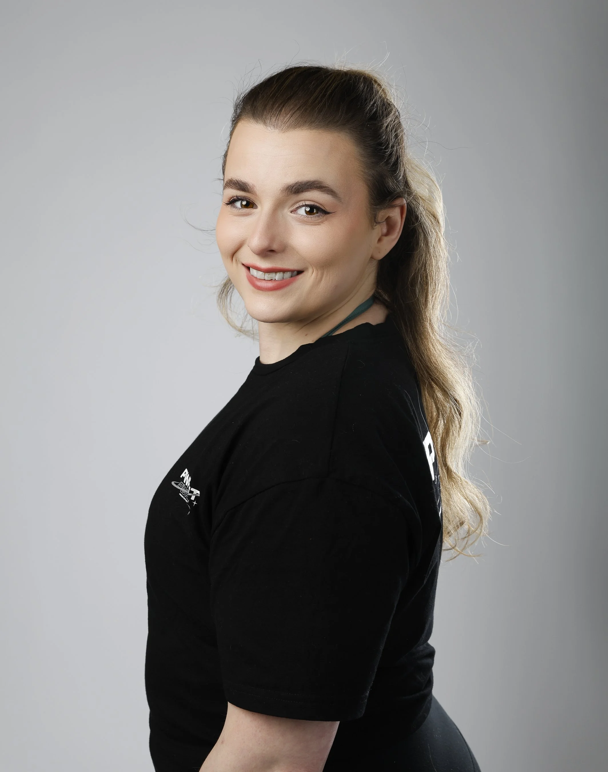 A young woman with long, wavy brown hair, and makeup, smiling at the camera. She is wearing a black shirt with white text and standing against a plain gray background.