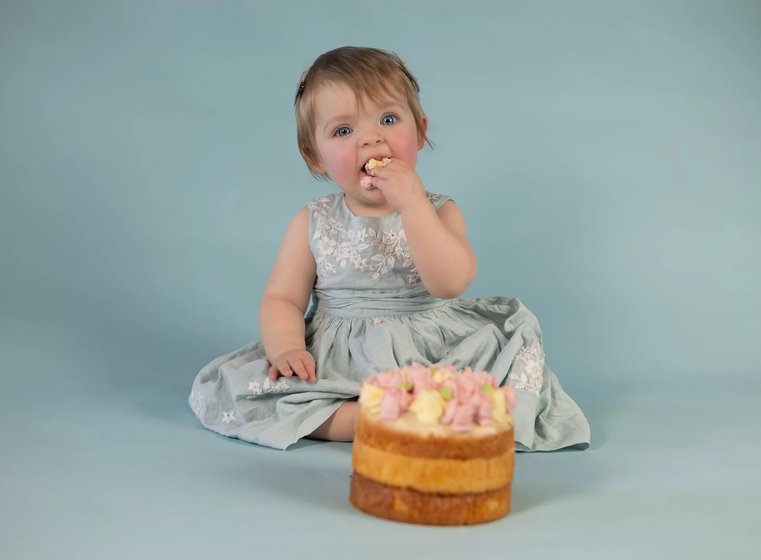 A young girl with blue eyes and light brown hair, wearing a gray dress with embroidered flowers, is sitting on a light blue background. She is eating something and has a surprised or curious expression. In front of her, there is a small layered cake 